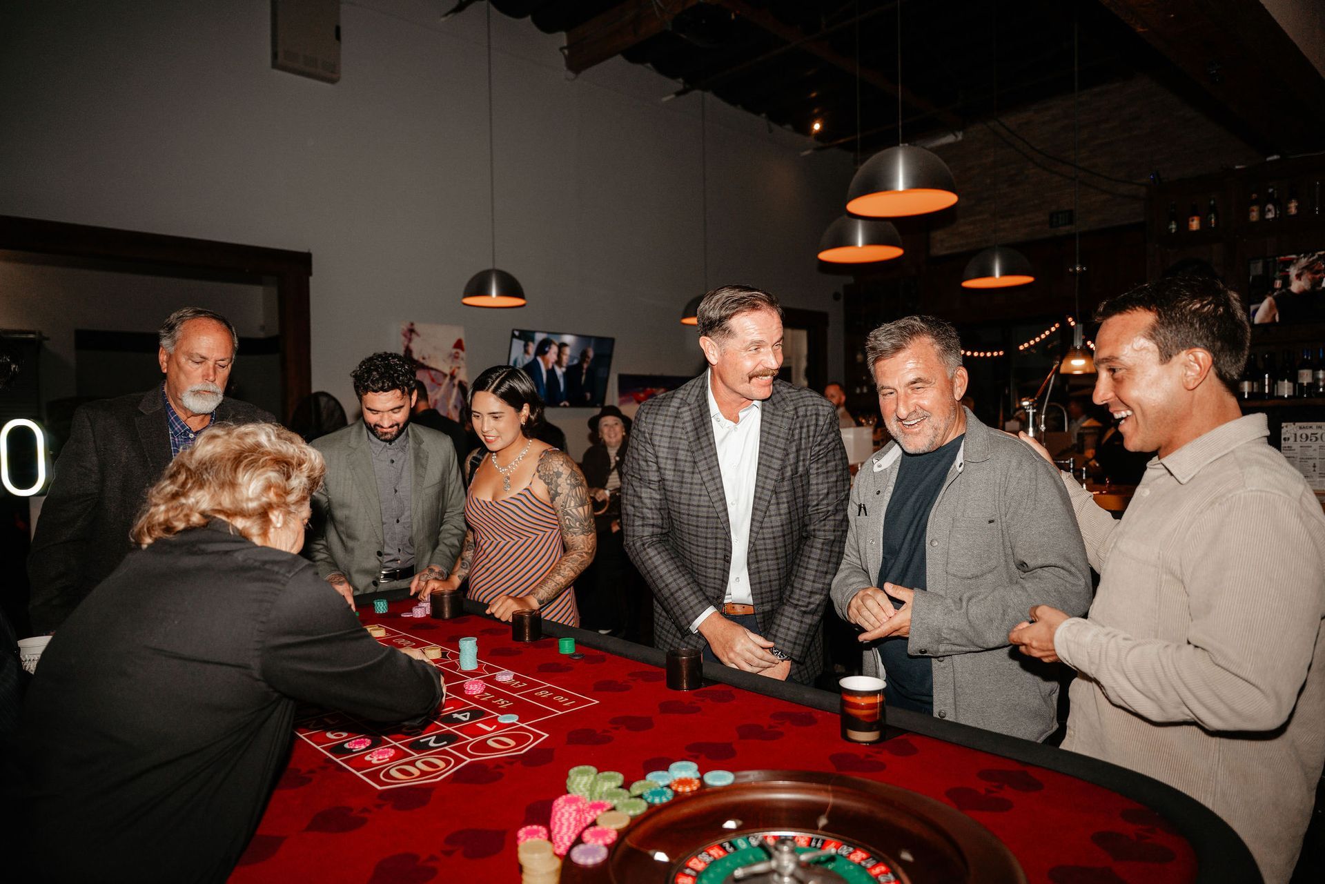 People gathered around a roulette table with red felt, chips, and a dealer in a dimly lit room.