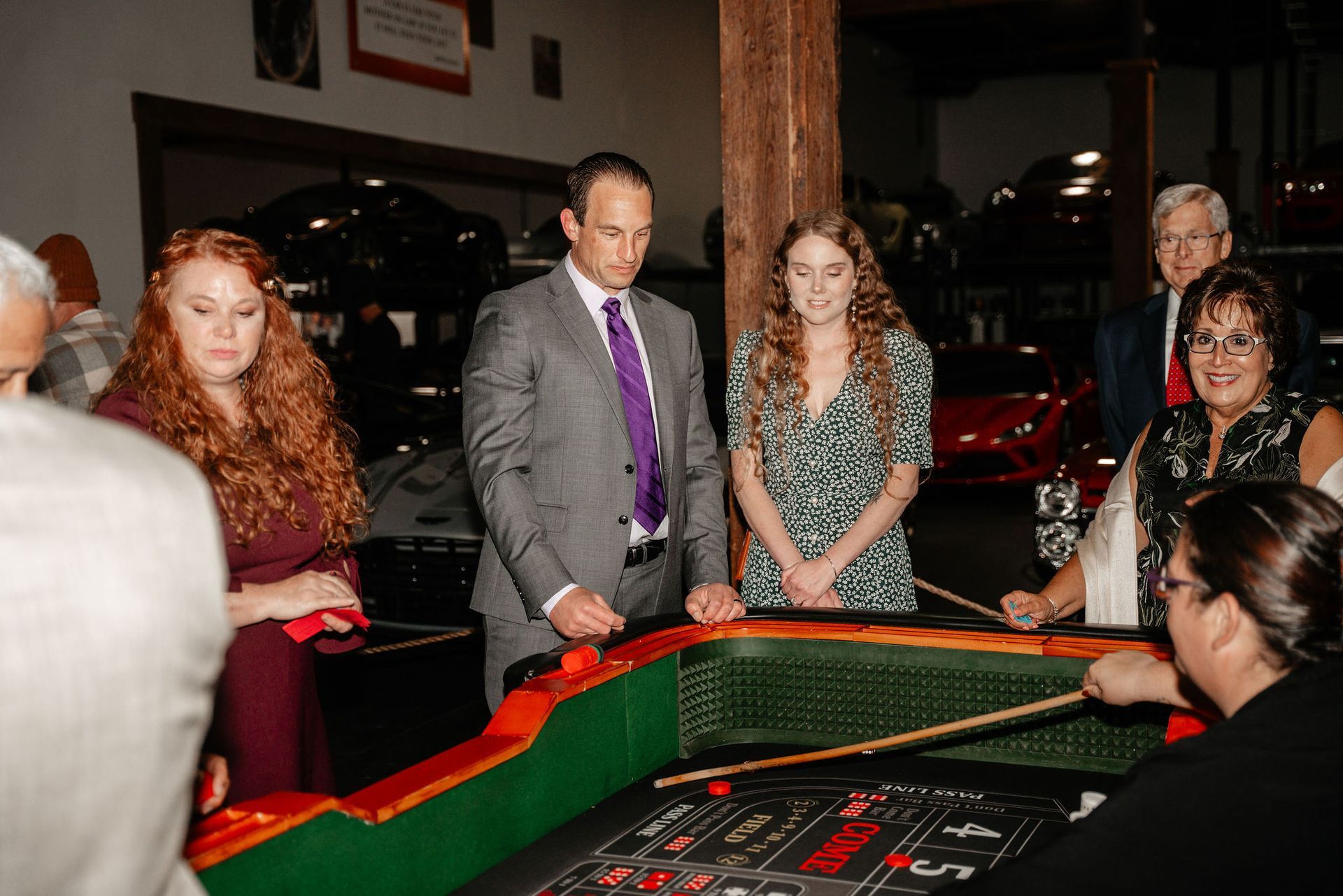 People gathered around a roulette table indoors. A man in a suit, a woman with red hair, and others watch the game. Cars are in the background.