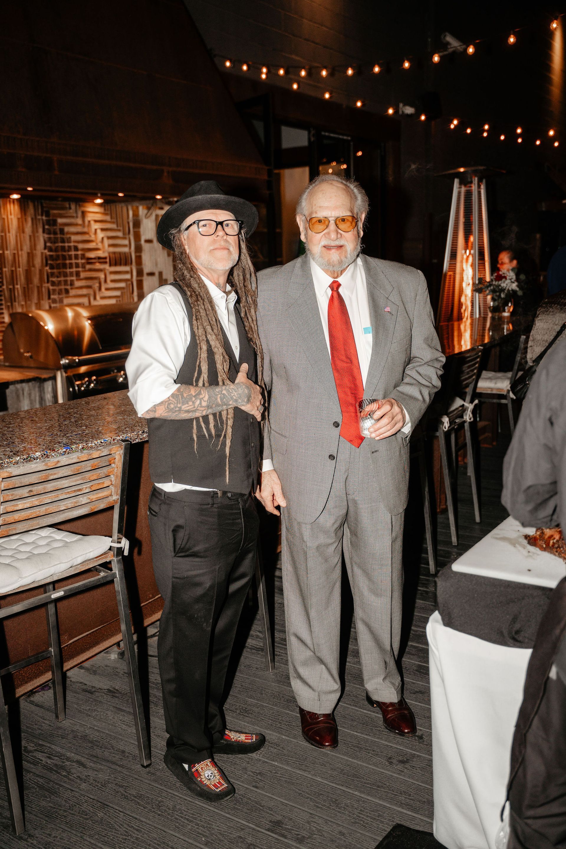 Two people, one in a hat and vest with dreadlocks, the other in a suit with a red tie, stand together at an event.