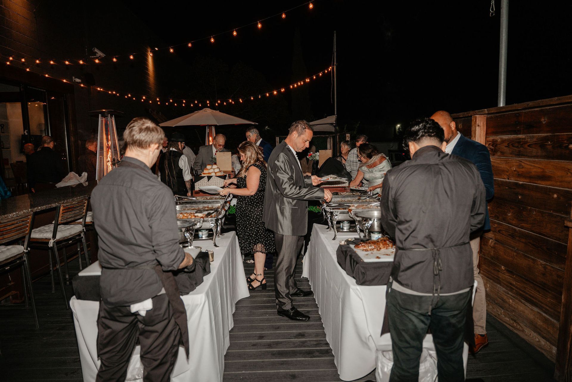 Buffet tables at an outdoor evening event. Guests serve food with staff nearby. String lights overhead.