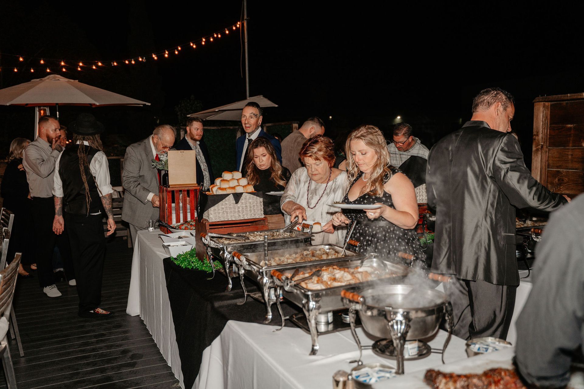 People at an outdoor buffet, serving themselves from steam tables on a white tablecloth at night.