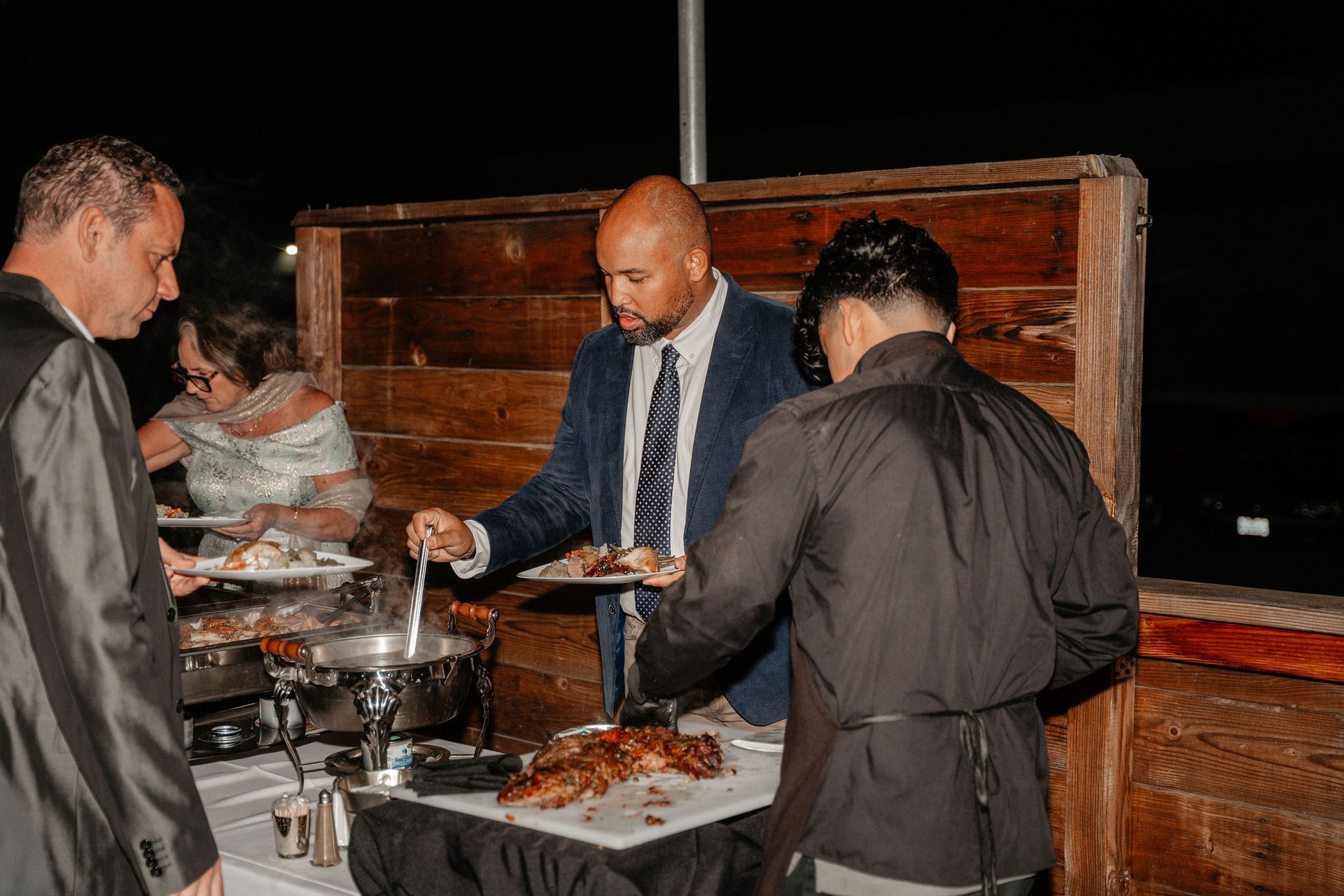 People serving themselves food from a buffet; man in suit ladling soup, another carving meat.
