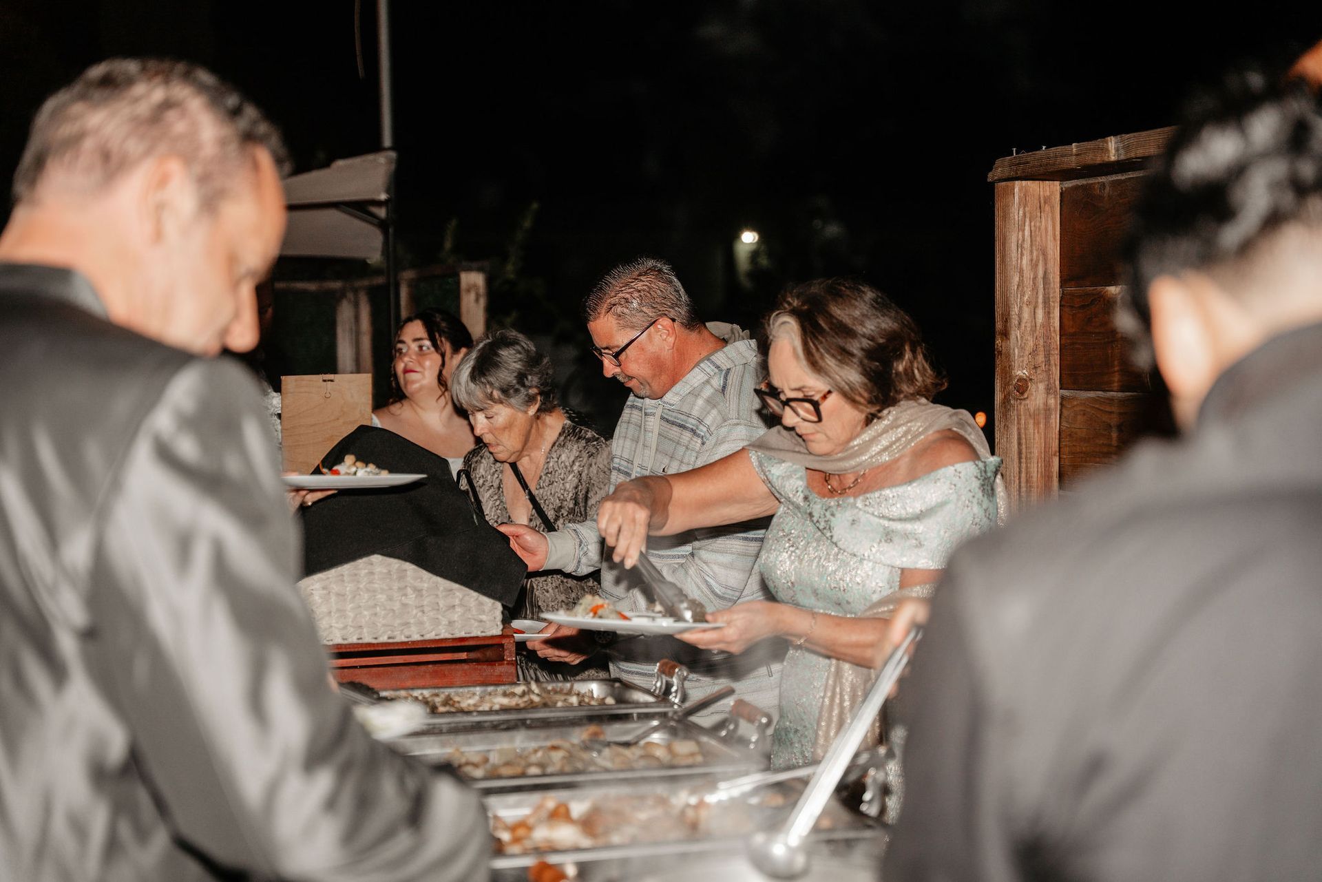 People serving themselves food at an outdoor buffet. Dark setting with a wooden structure and serving tables.