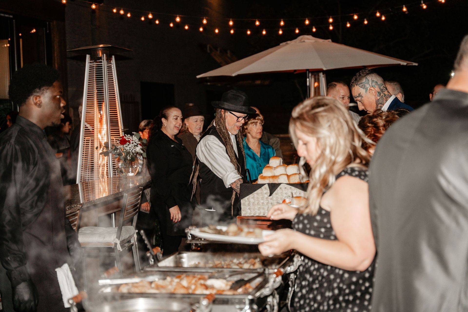 People at an outdoor catering buffet, food served from stainless steel trays, string lights overhead.