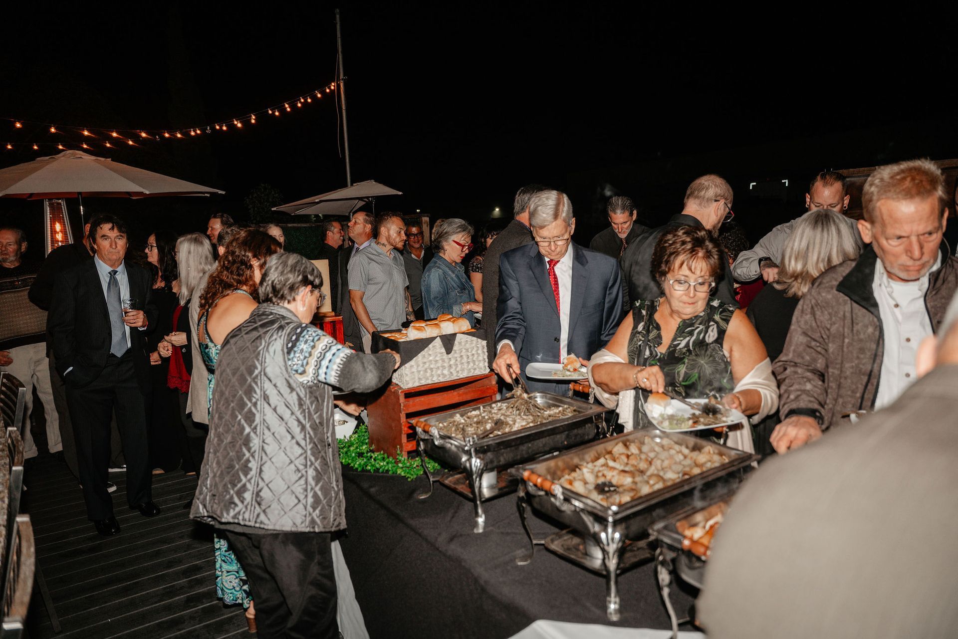People at an outdoor buffet, serving themselves from warming trays at night. String lights overhead.