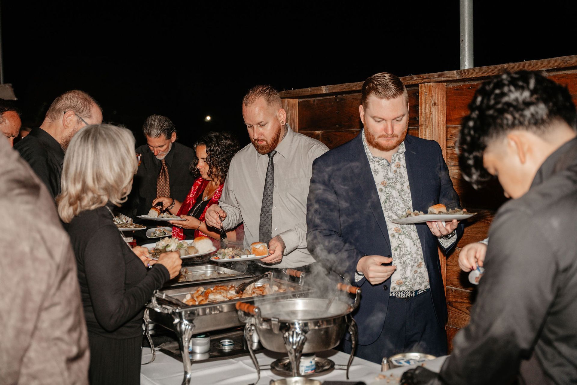 People serving themselves food from a buffet table at an event, outdoors at night.