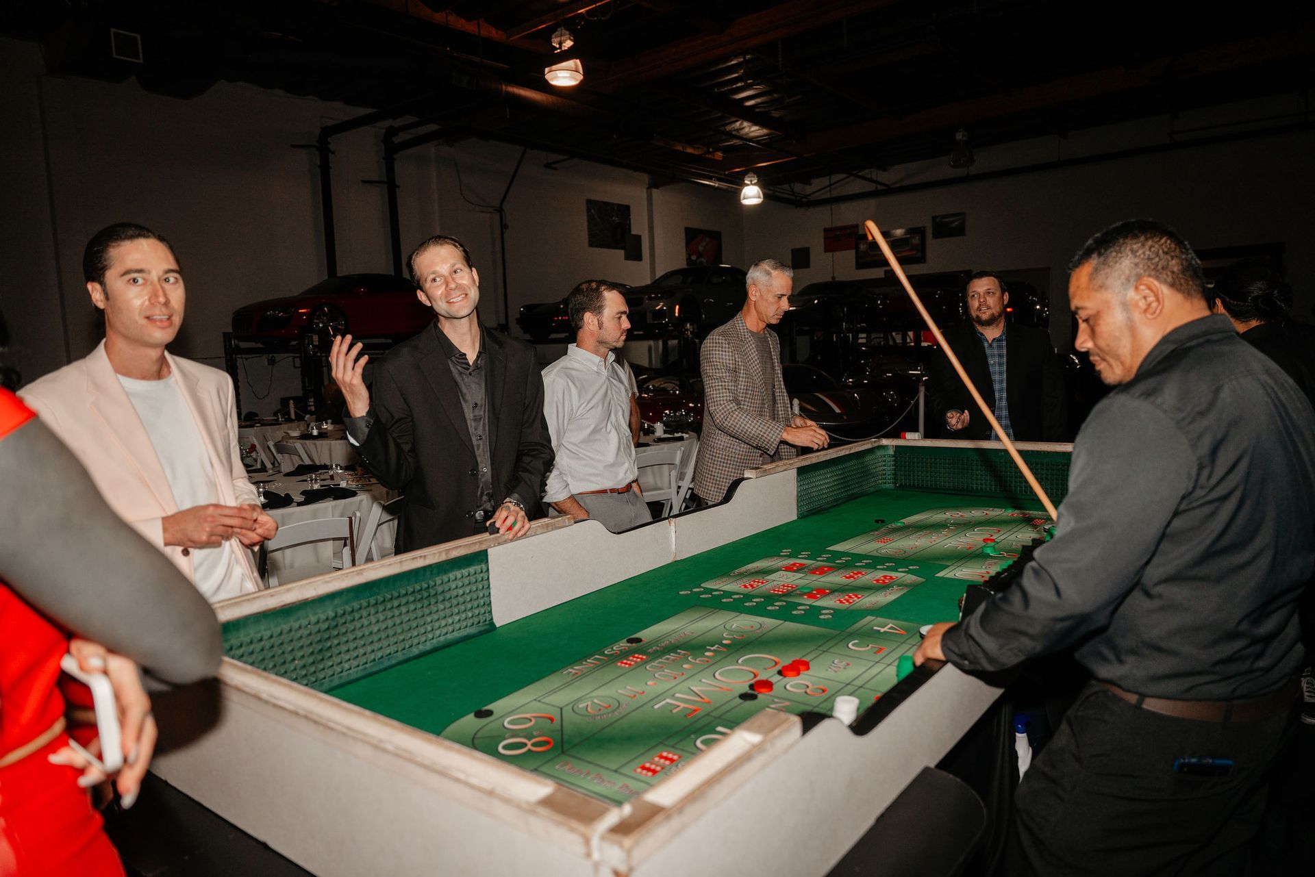 People playing a tabletop game in a garage. Green playing surface, men using cues, cars in background.