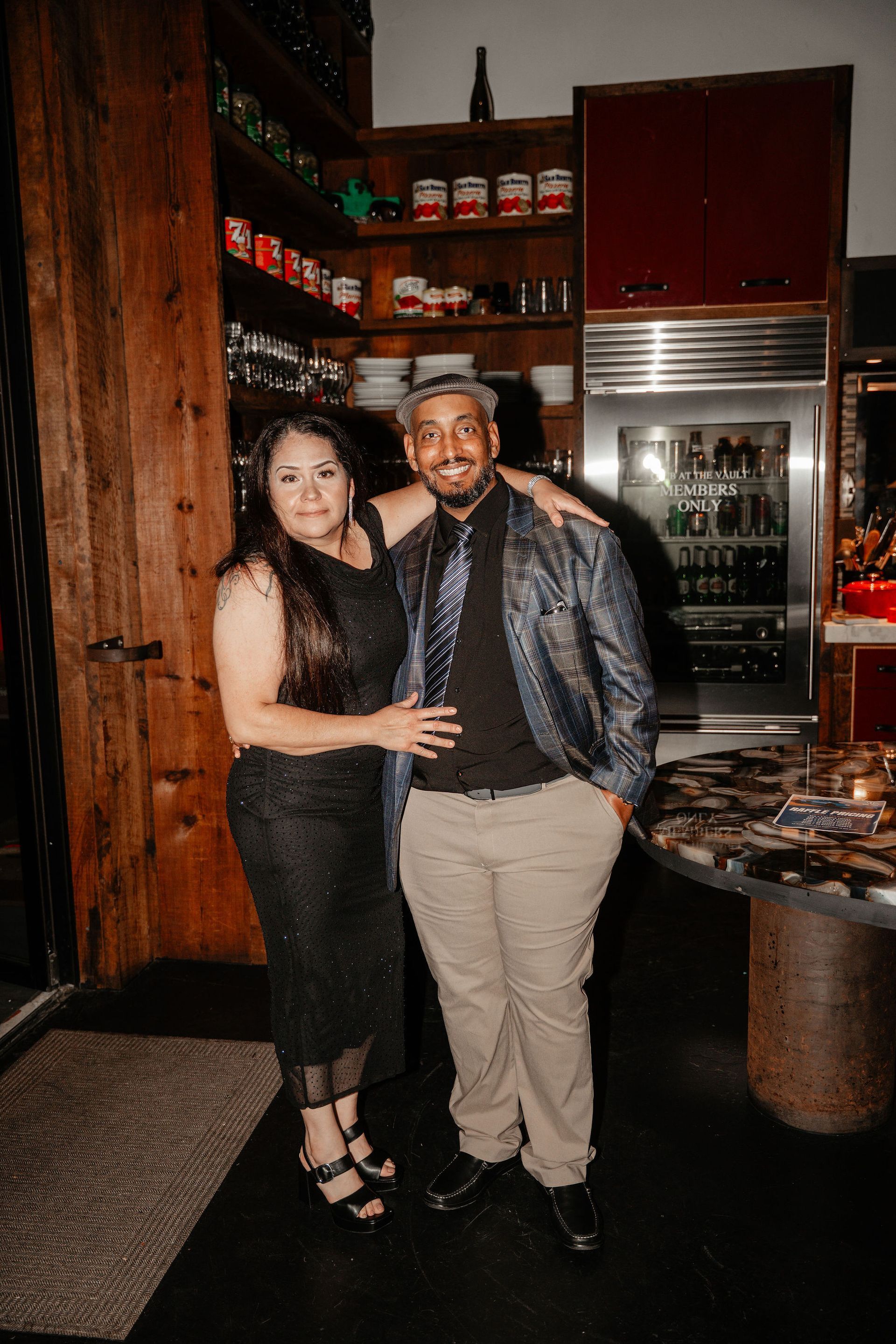 Couple posing indoors; woman in black dress, man in sequined jacket, by wooden door.