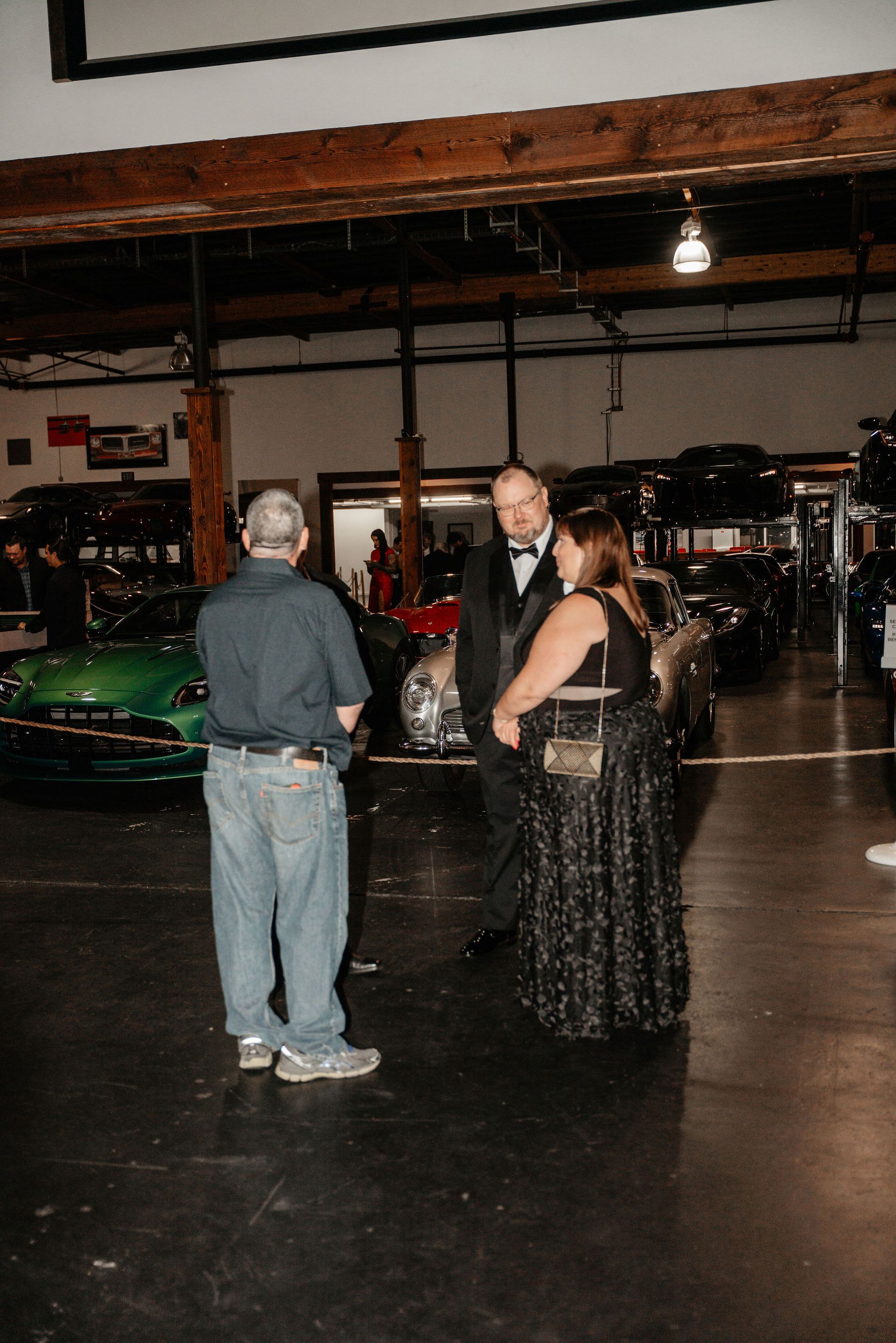 People conversing in a garage among classic cars; one in tuxedo, one in a gown, and another in casual attire.