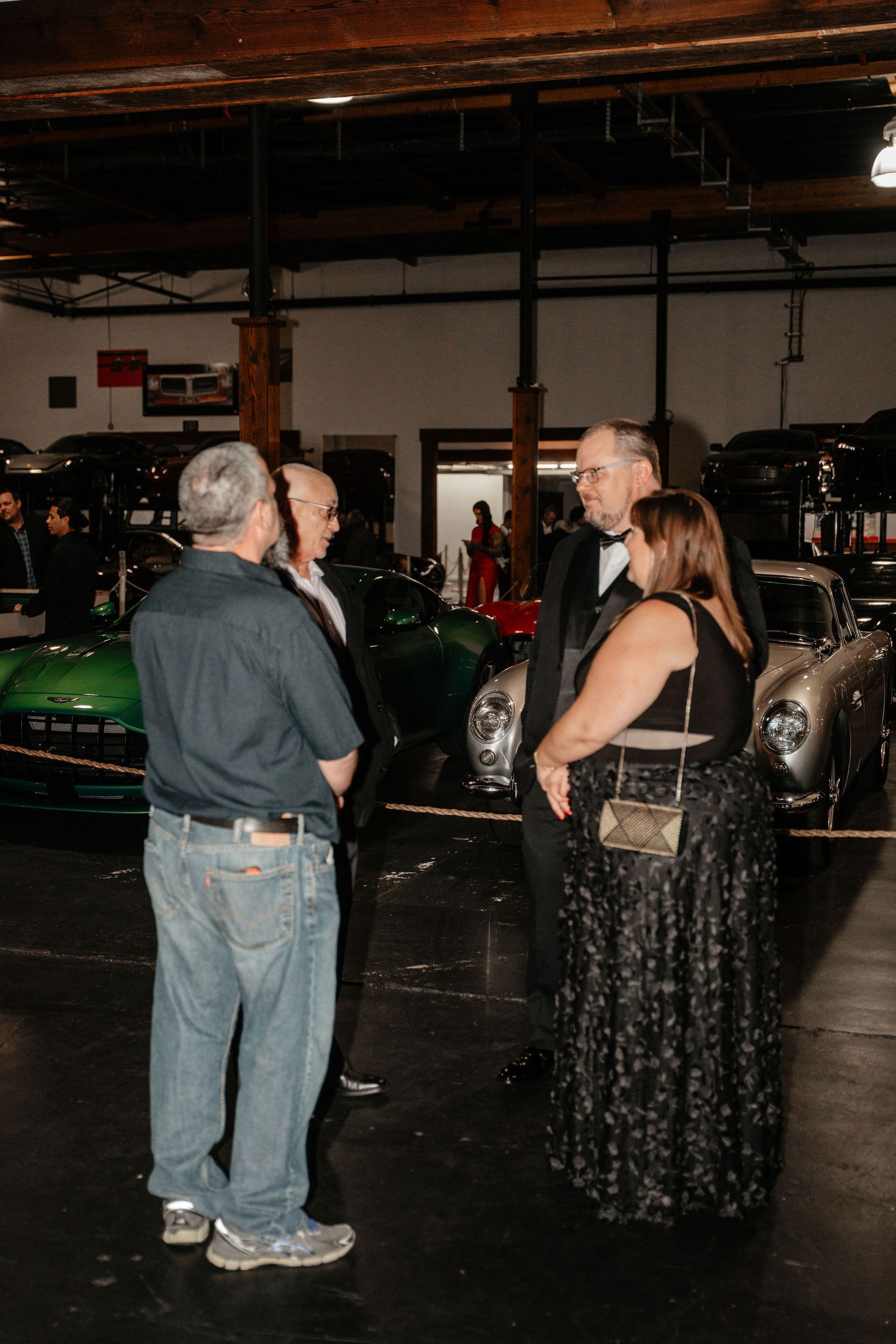 Four people in formal attire converse near classic cars in a garage setting.