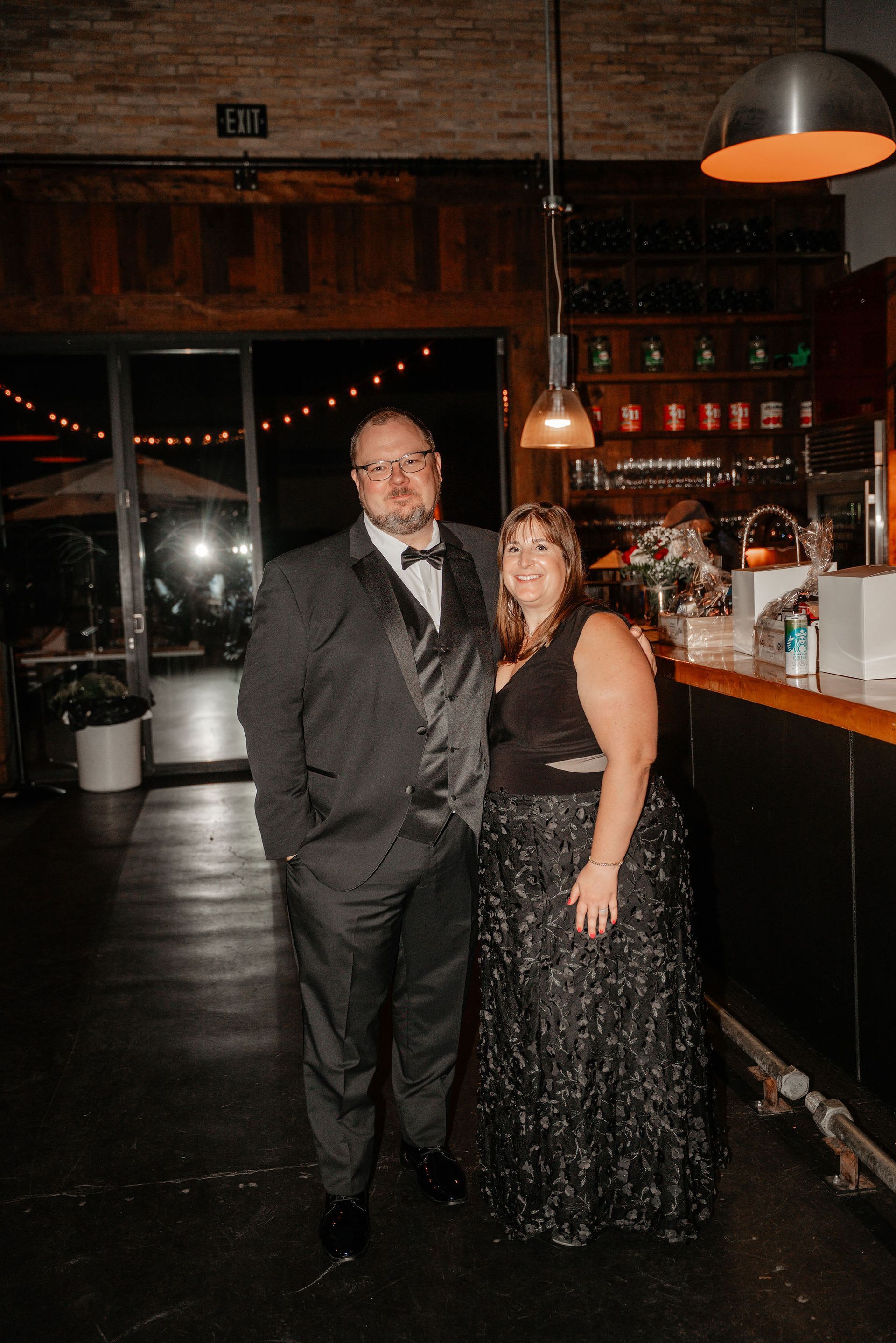 Couple in formal attire at an event, man in a tuxedo, woman in a black gown, standing near a bar.