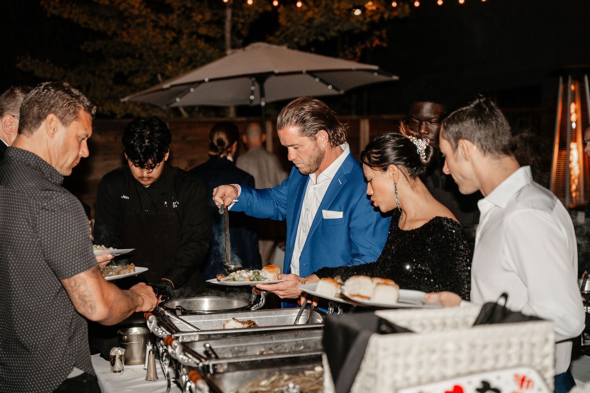 People serving themselves at a buffet table, outdoors at night. Man in blue blazer ladles food. Other guests reach for food.
