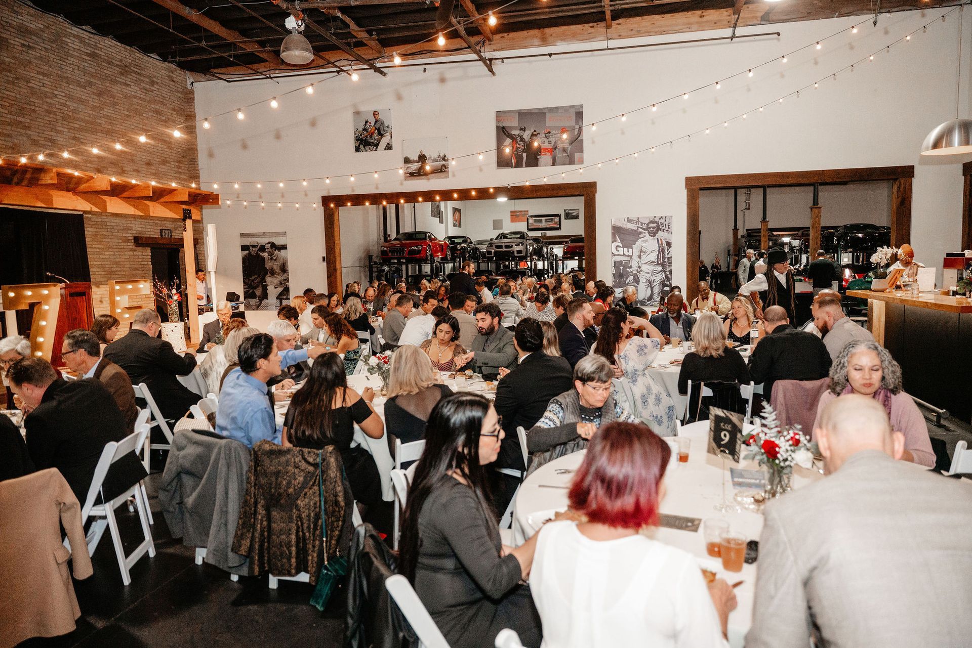Large gathering at tables under string lights in an industrial-style event space. People are seated and talking.