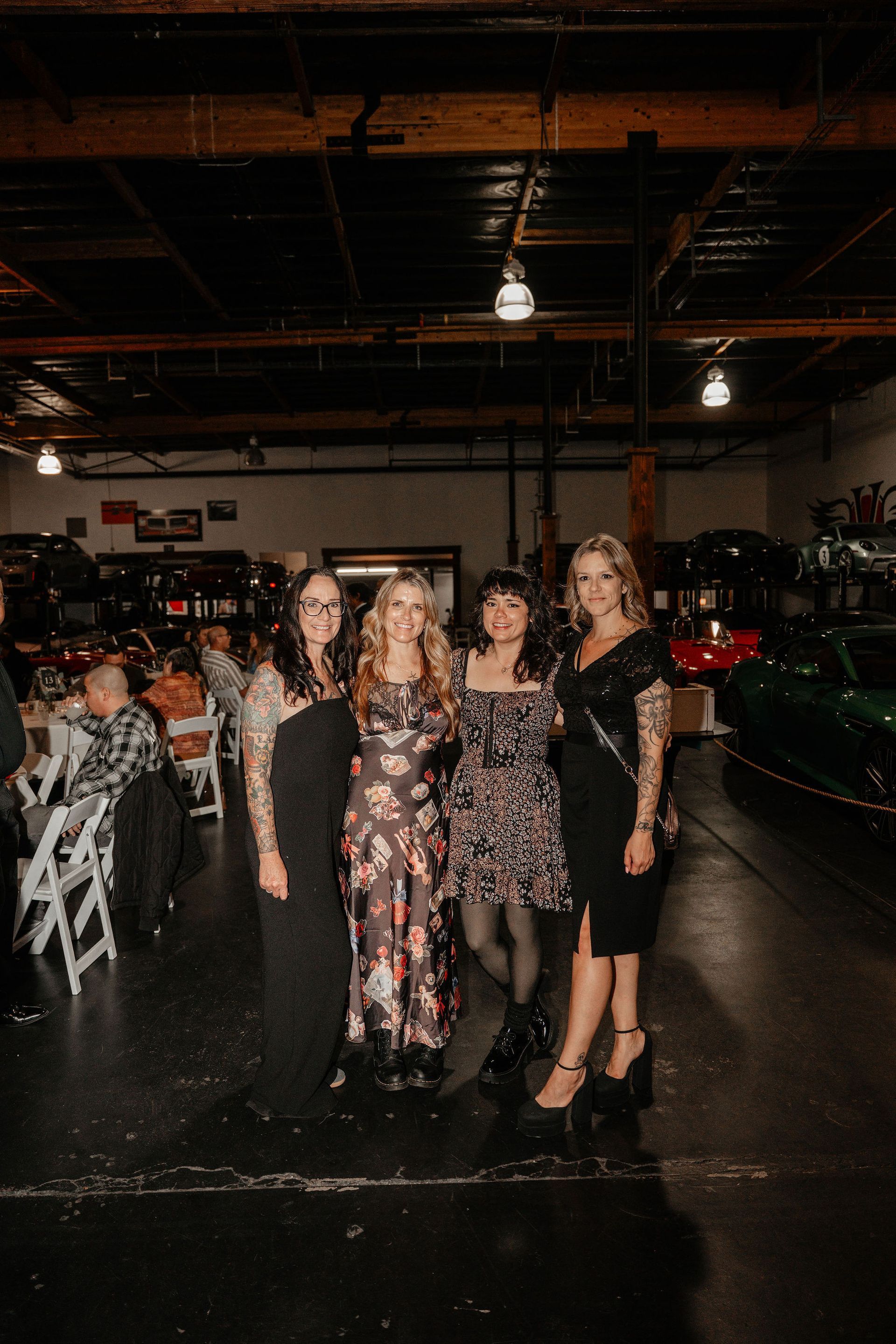 Four women pose in a garage with classic cars. They wear dresses, smiling, under overhead lights.