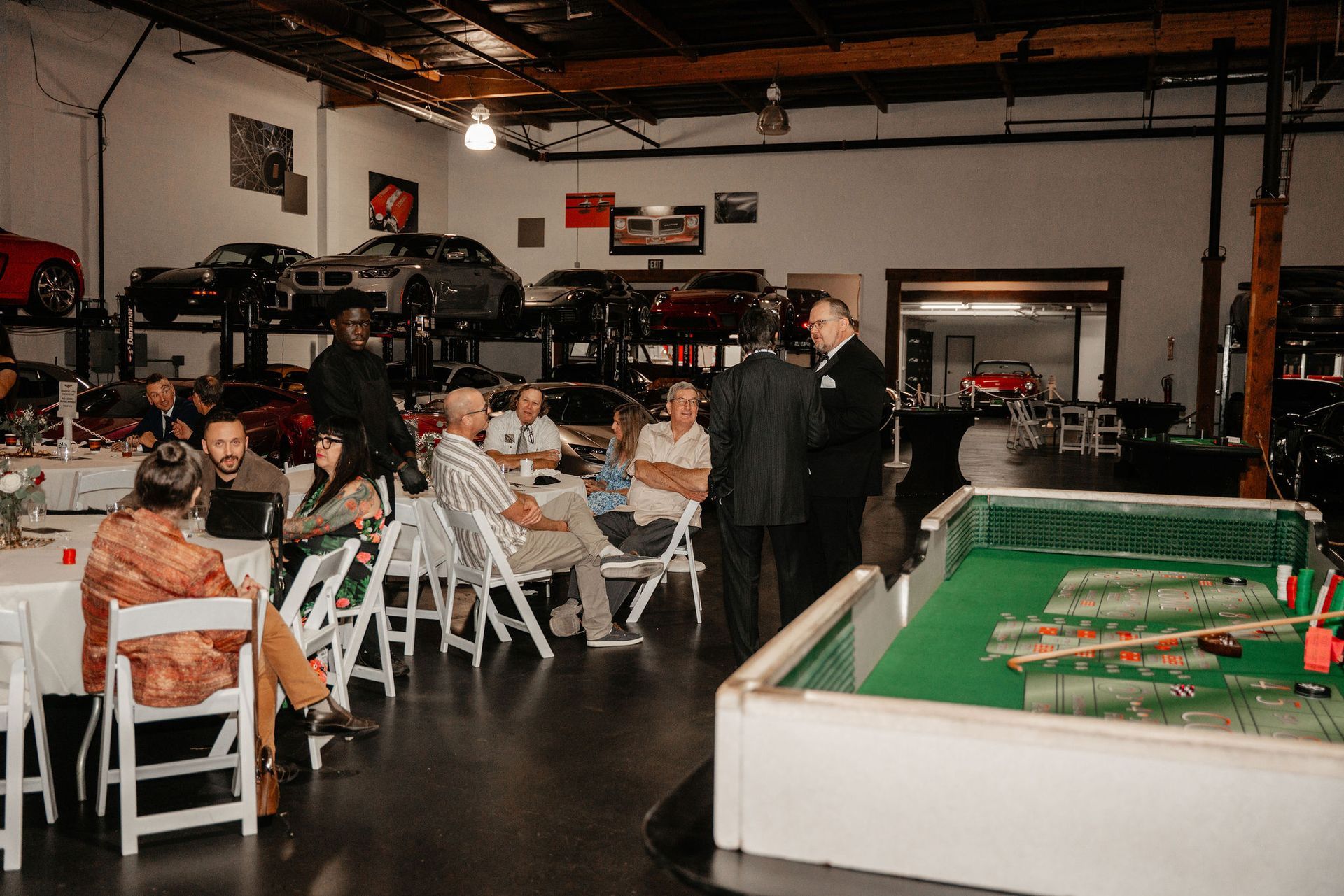 People gathered in a garage-like space, some seated at tables. Cars are visible on lifts and throughout the room.