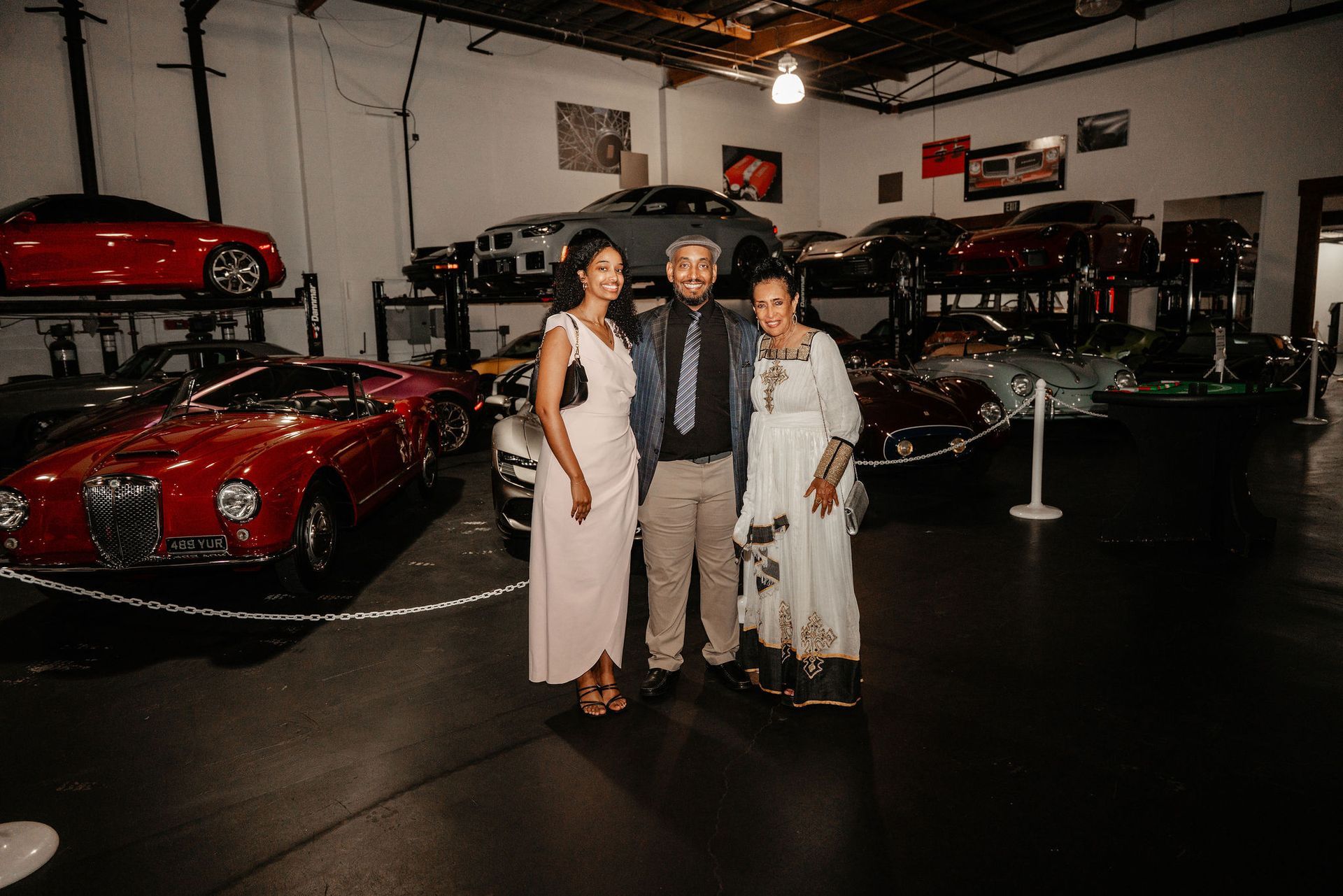 Three people pose in a car garage with various luxury cars.