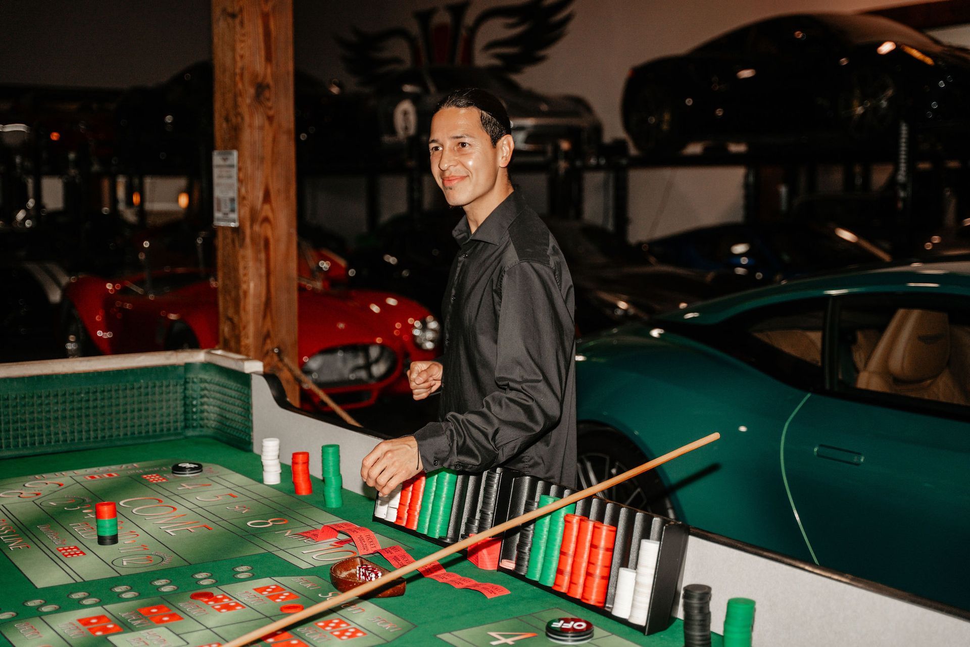 Man smiles at a craps table, surrounded by luxury cars in a garage setting.