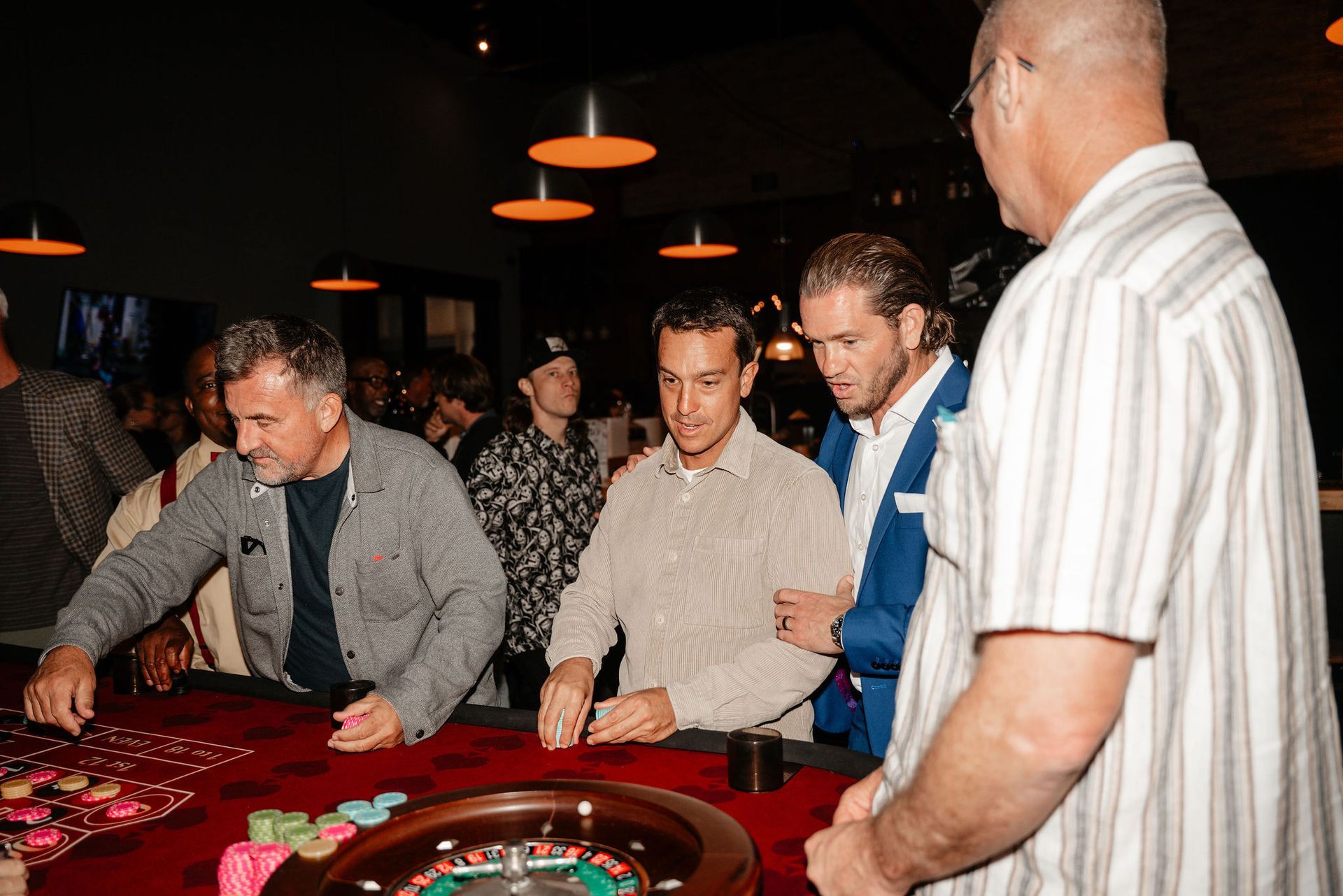 People at a casino table; several men placing chips, roulette wheel visible, low lighting.