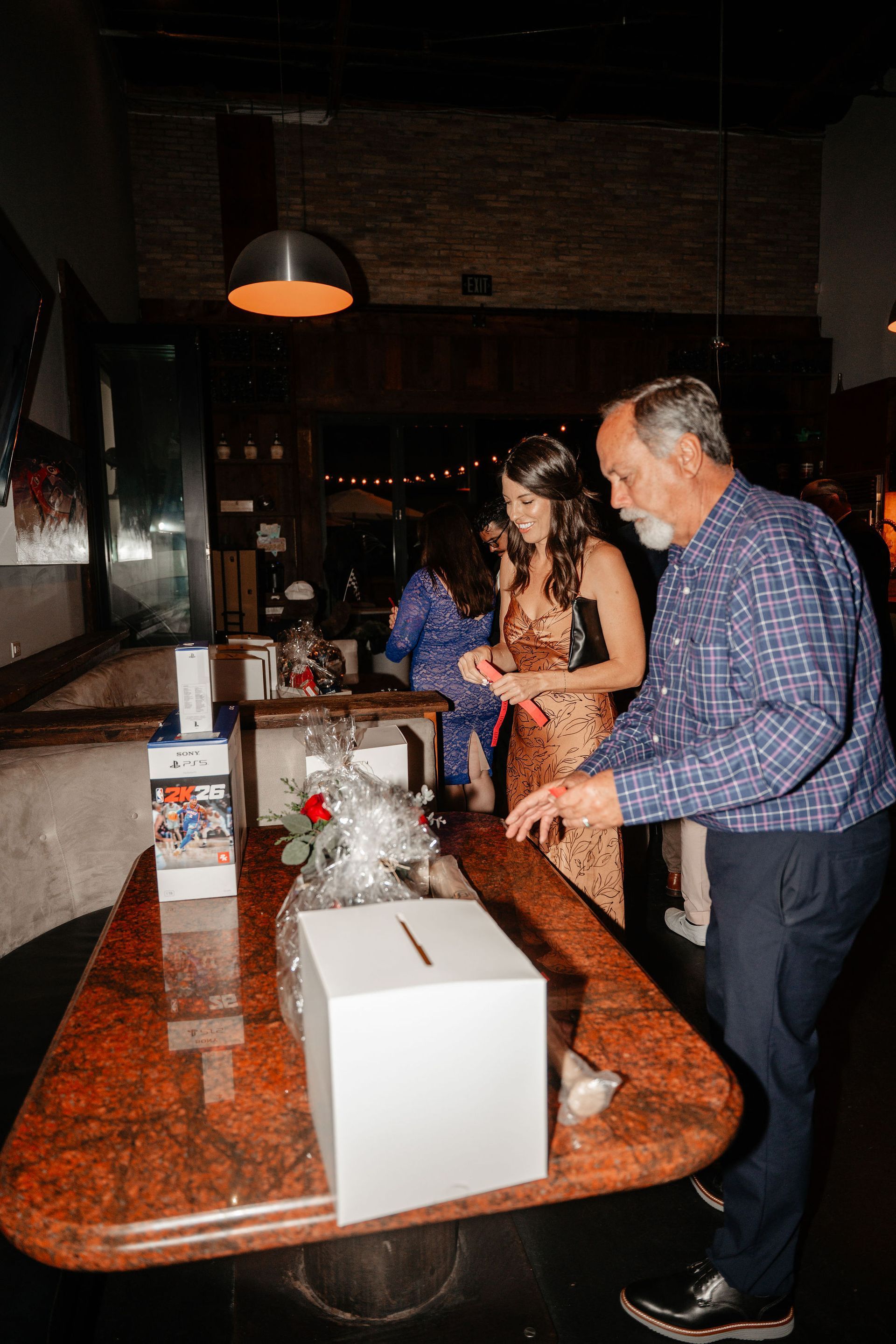 People near a table with a donation box and gift. A man reaches, a woman looks on, indoors.