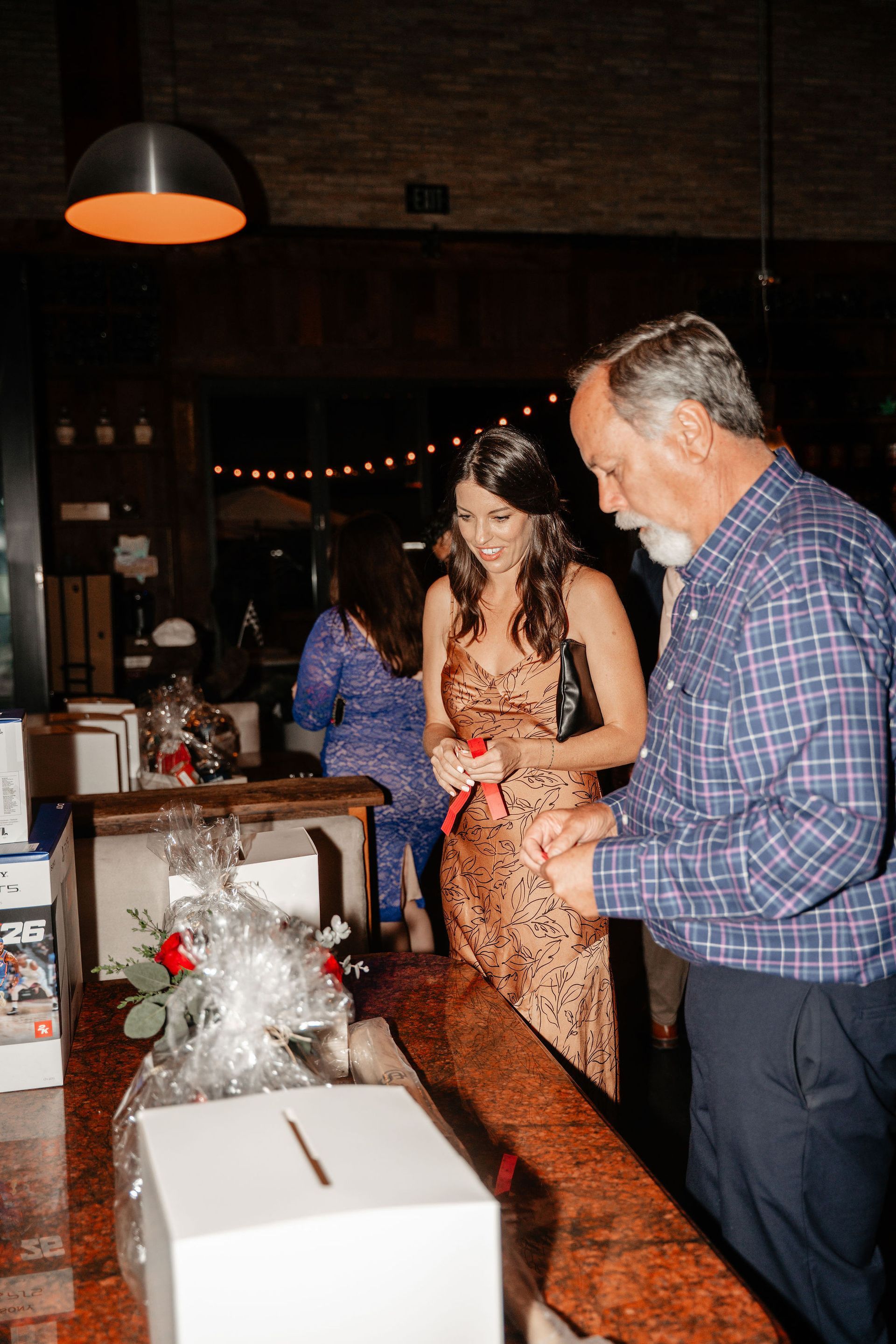 Woman and man at an event, near gift baskets, opening ribbon. Dimly lit bar.
