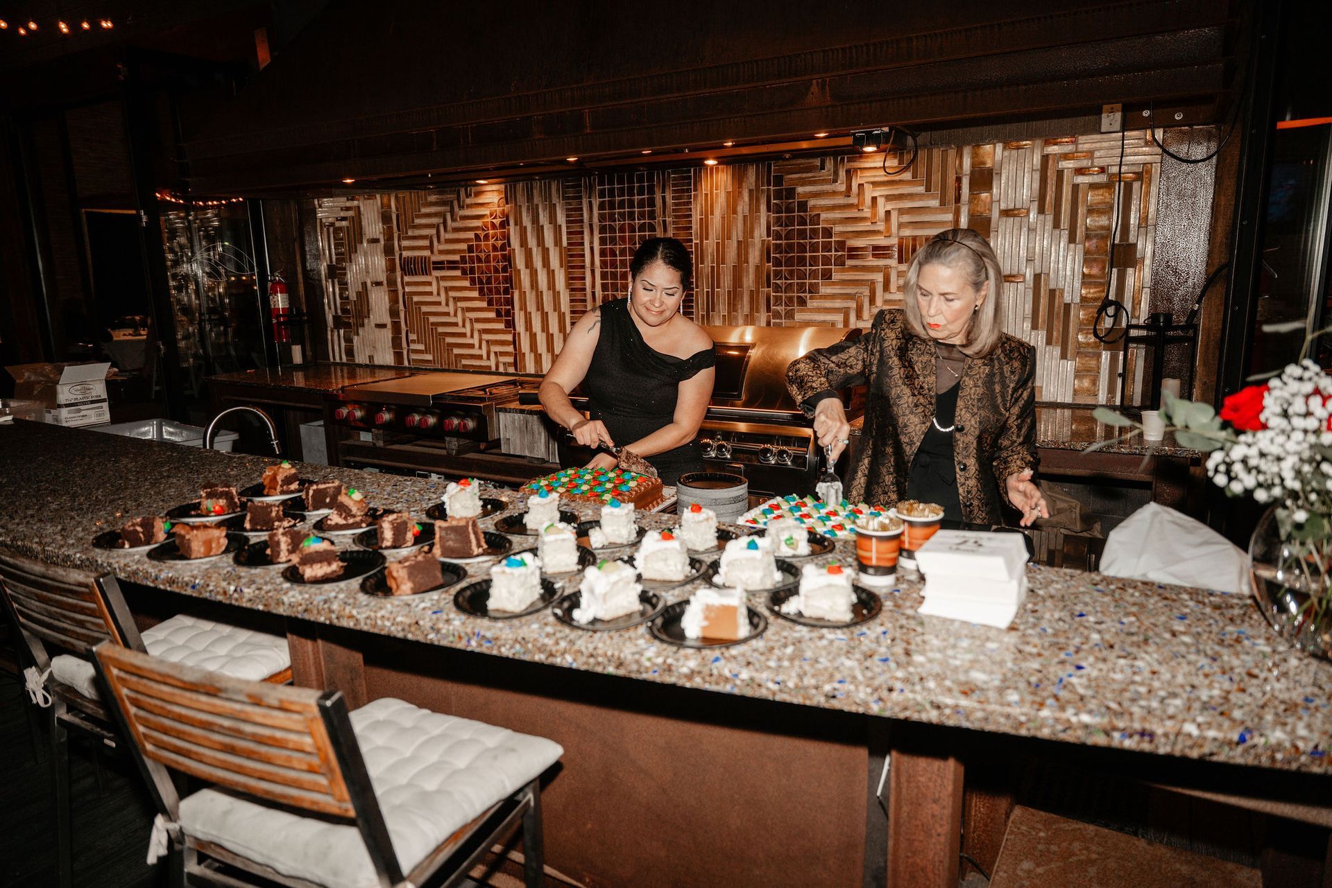 Two people preparing desserts behind a bar. Various desserts are displayed on plates. The bar has a textured countertop.