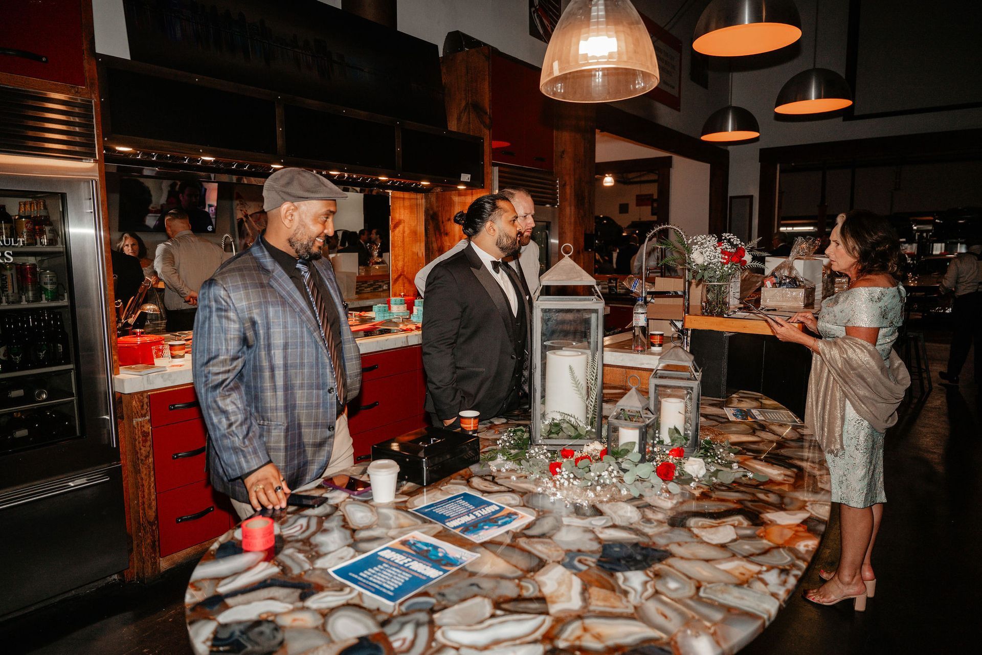 People at a decorated table, speaking. Candles, flowers, and cards are displayed. Dark room with overhead lights.
