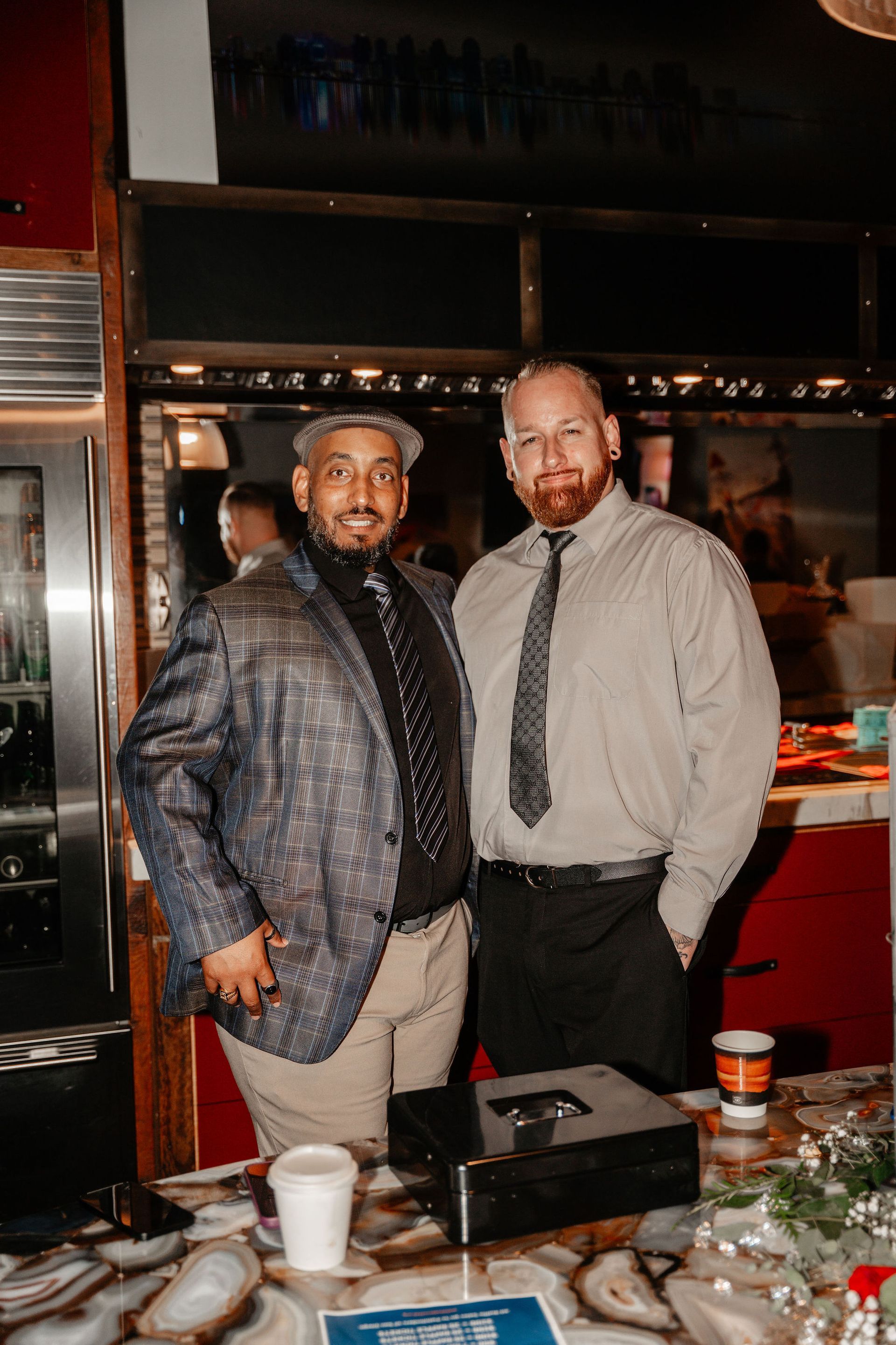 Two men smiling, one in a patterned blazer and hat, the other in a button-down shirt, standing indoors.