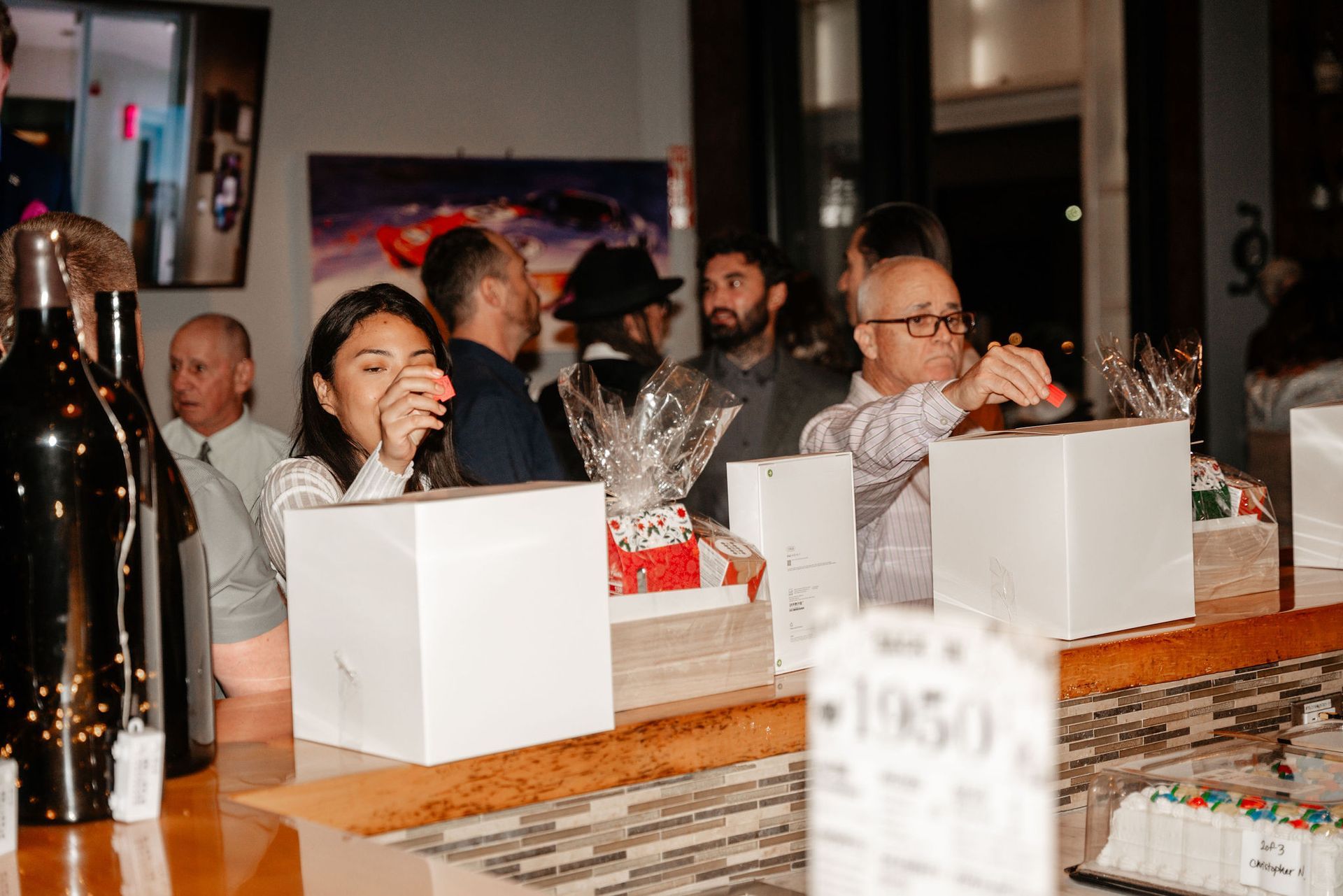 People at a bar, some reach for items on the counter. A woman touches her face. White boxes and gifts.