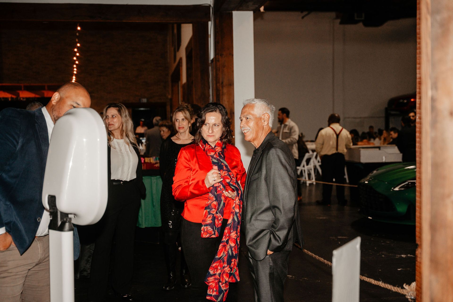 People gathered at an event entrance; a woman in red jacket talks to an older man, a photo booth in front.