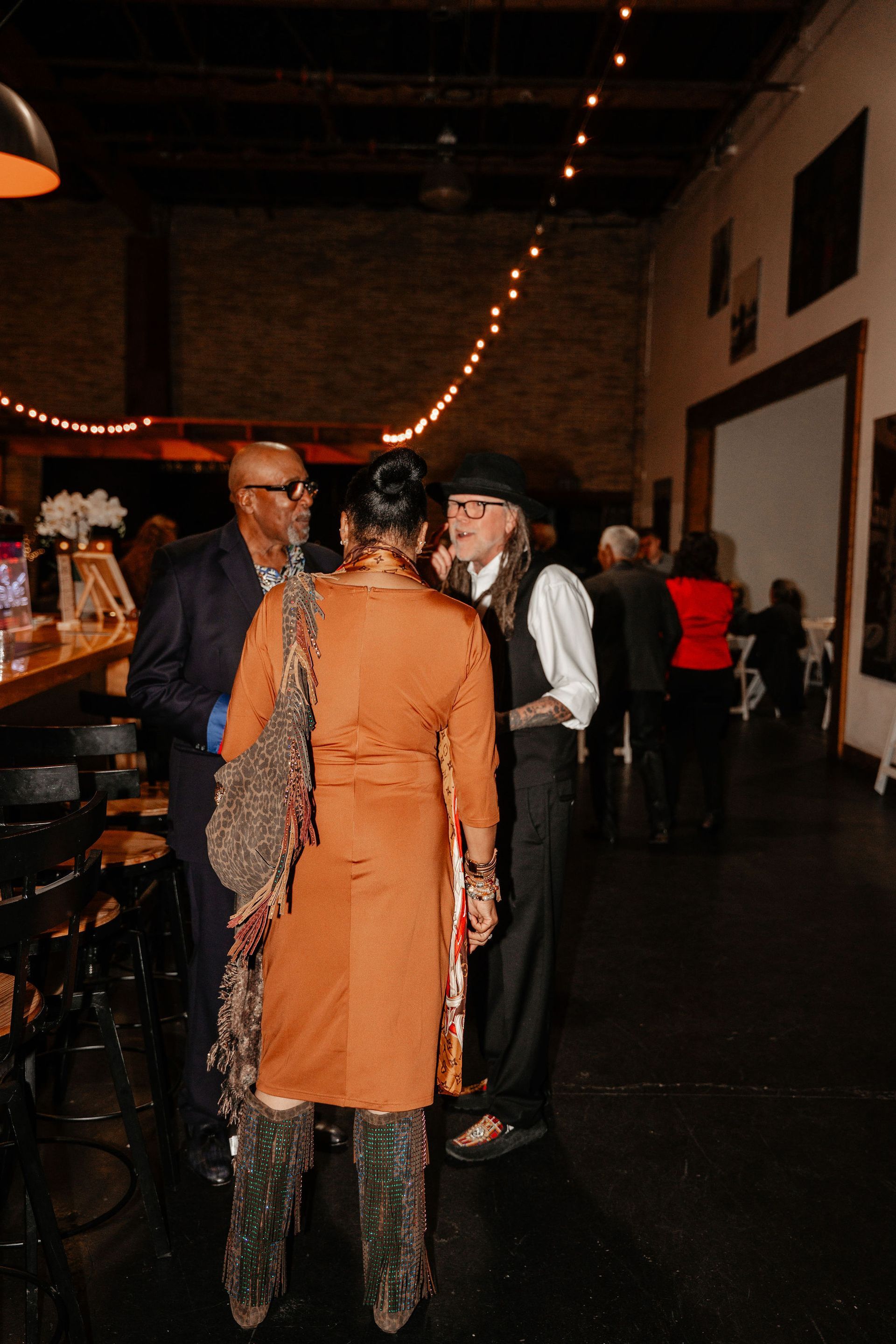 People conversing at an event in a dimly lit industrial space; one wears a suit, another a long coat.