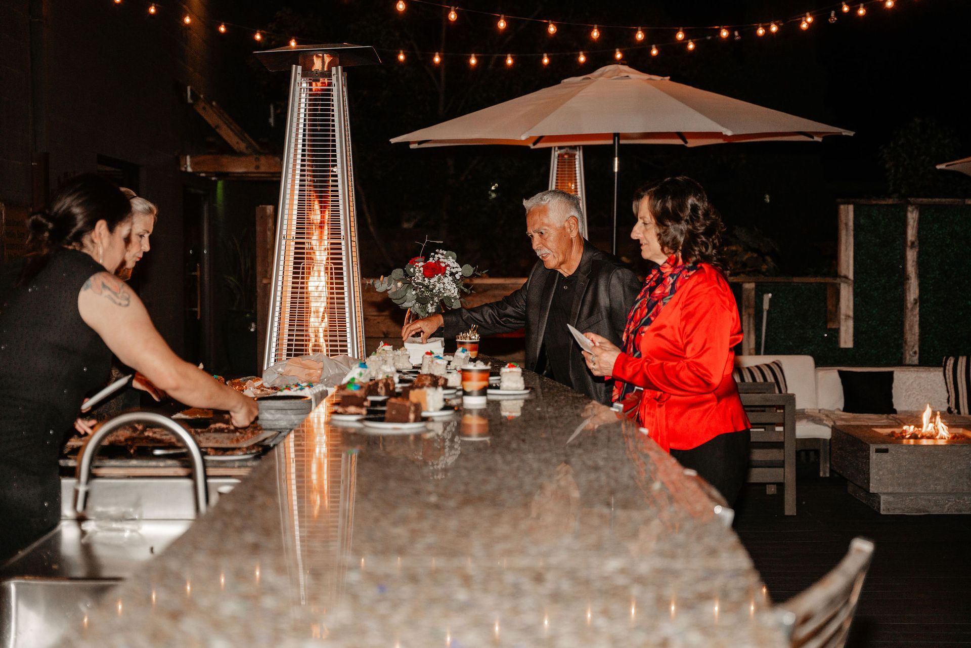People at an outdoor bar at night. A bartender serves drinks. String lights and a heater are in the background.