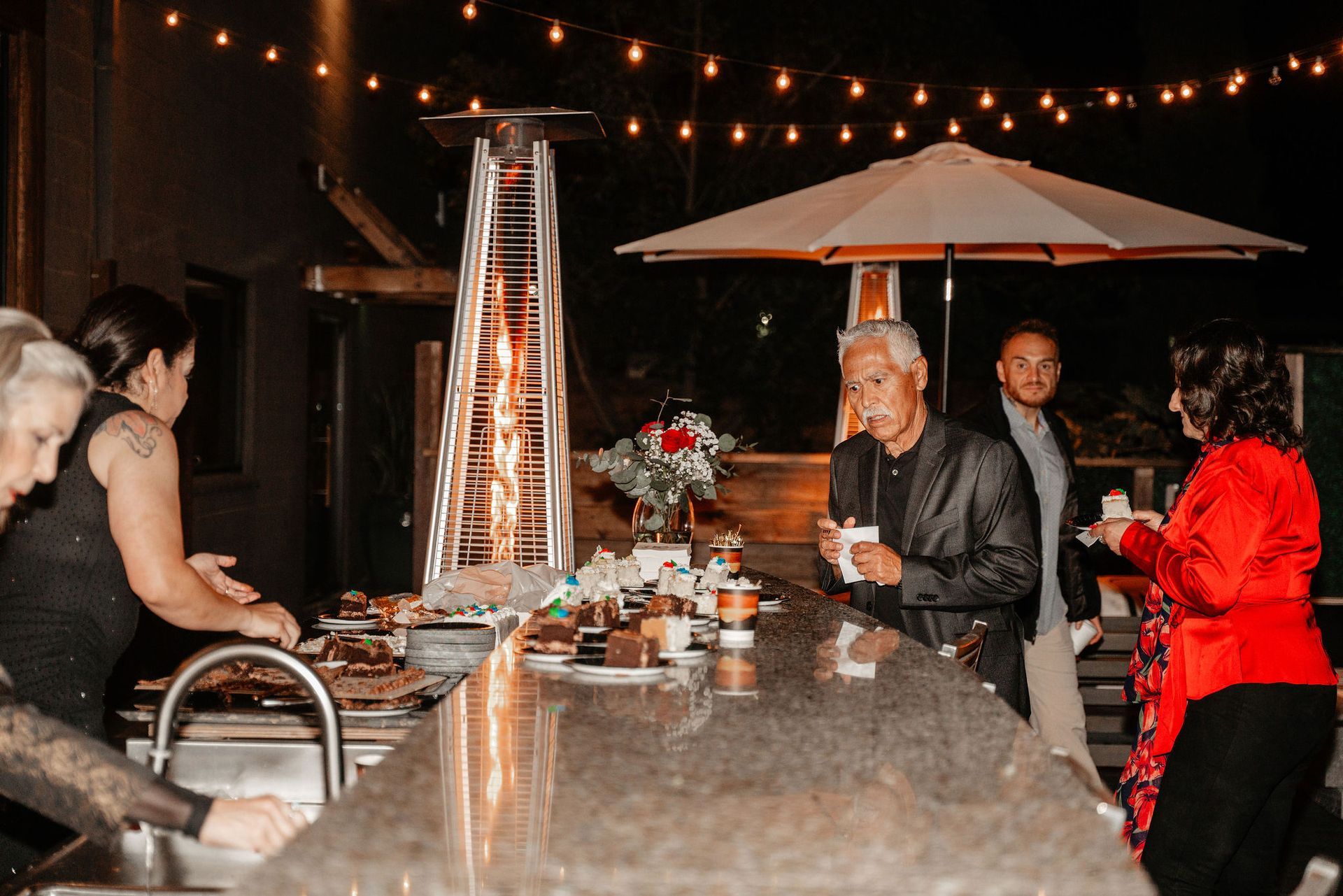 People at an outdoor party at night around a bar, with food and a tall heater.
