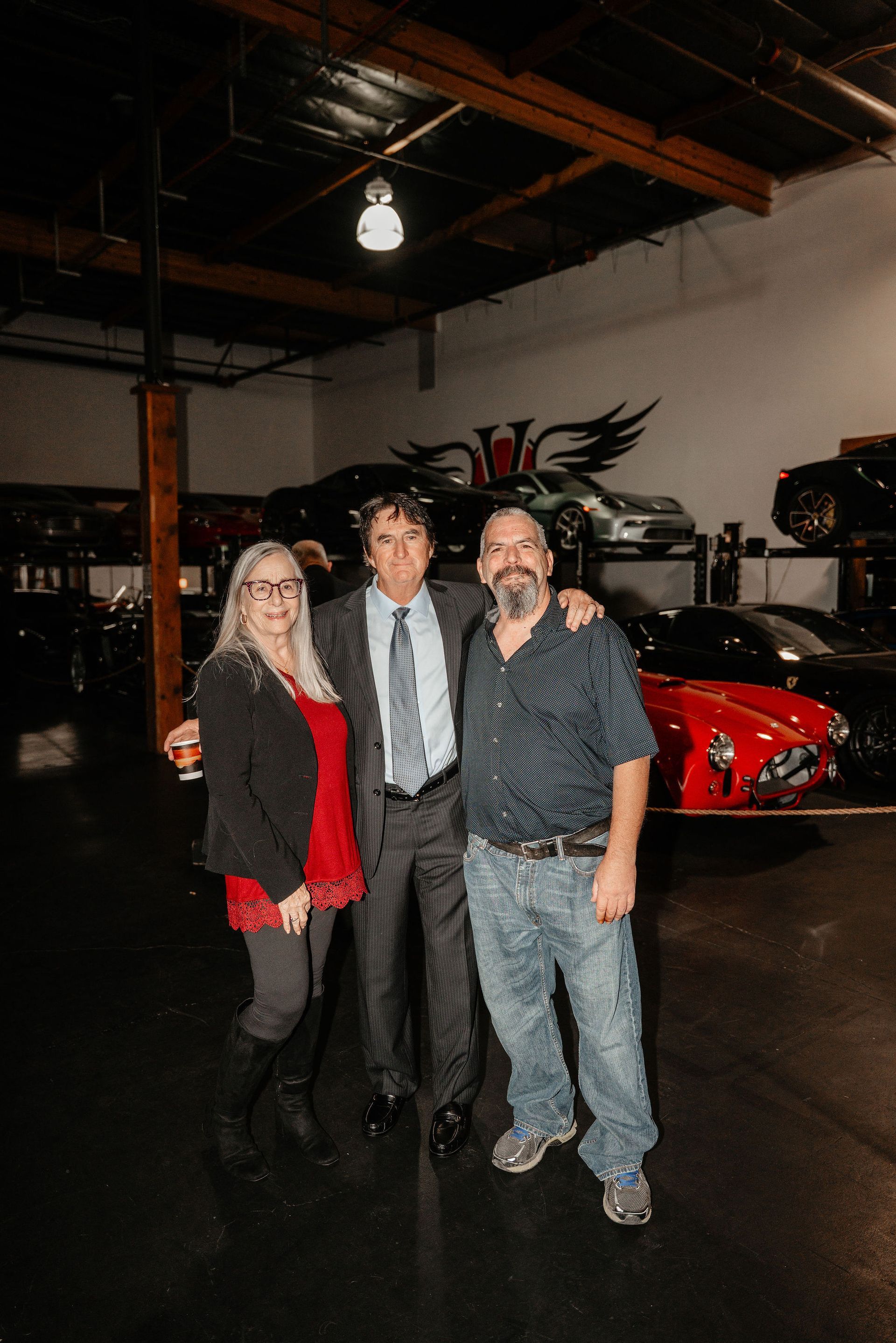 Three people smiling in a car repair shop; two men have their arms around each other. A red sports car is visible.