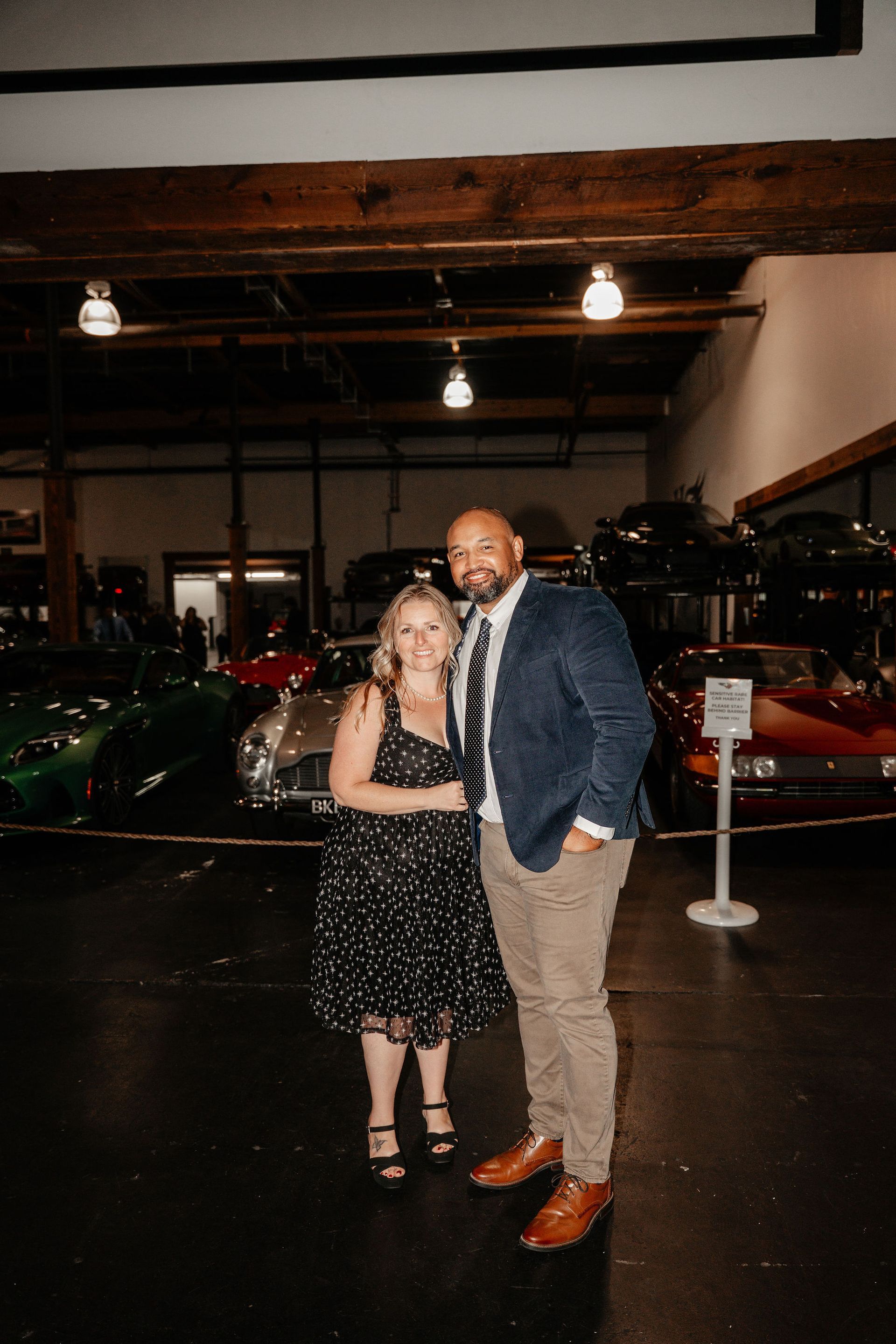 Couple posing in front of vintage cars. Woman in black dress, man in blue jacket and khaki pants. Indoor setting.