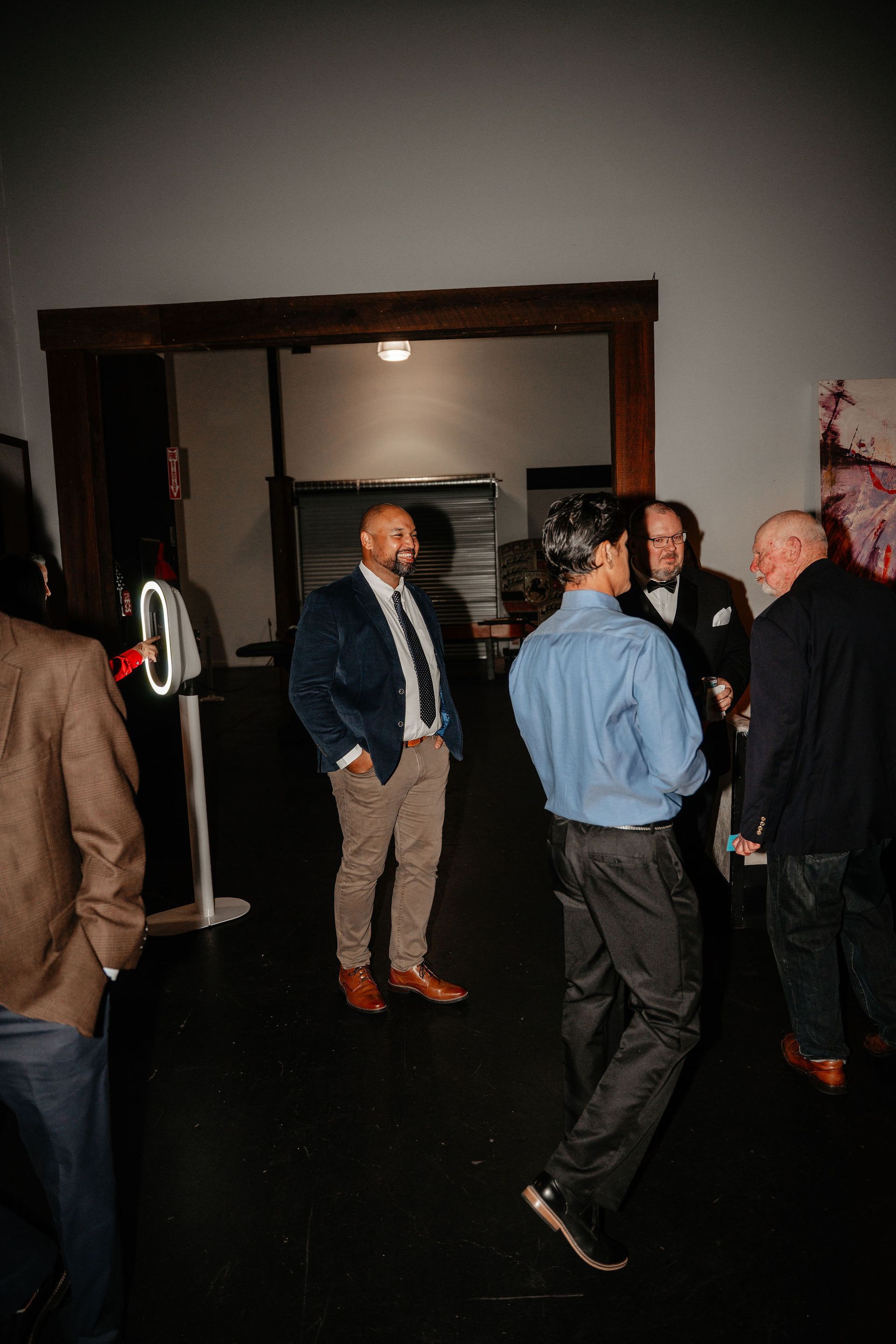 People in a room, some in suits, others in casual wear, talking, near an open doorway.