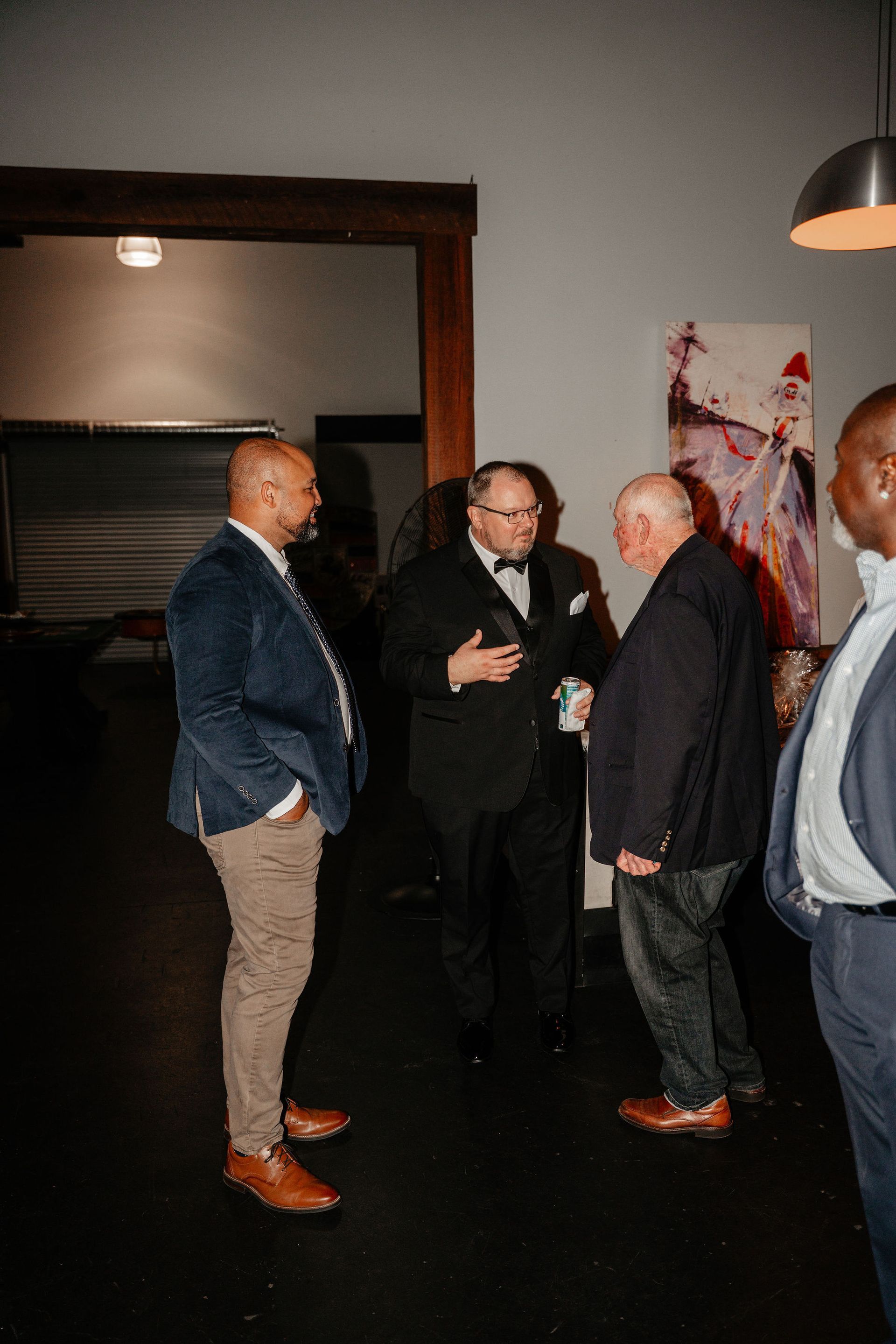 Men in a dimly lit room, some in suits, conversing near a painting and exposed beam.