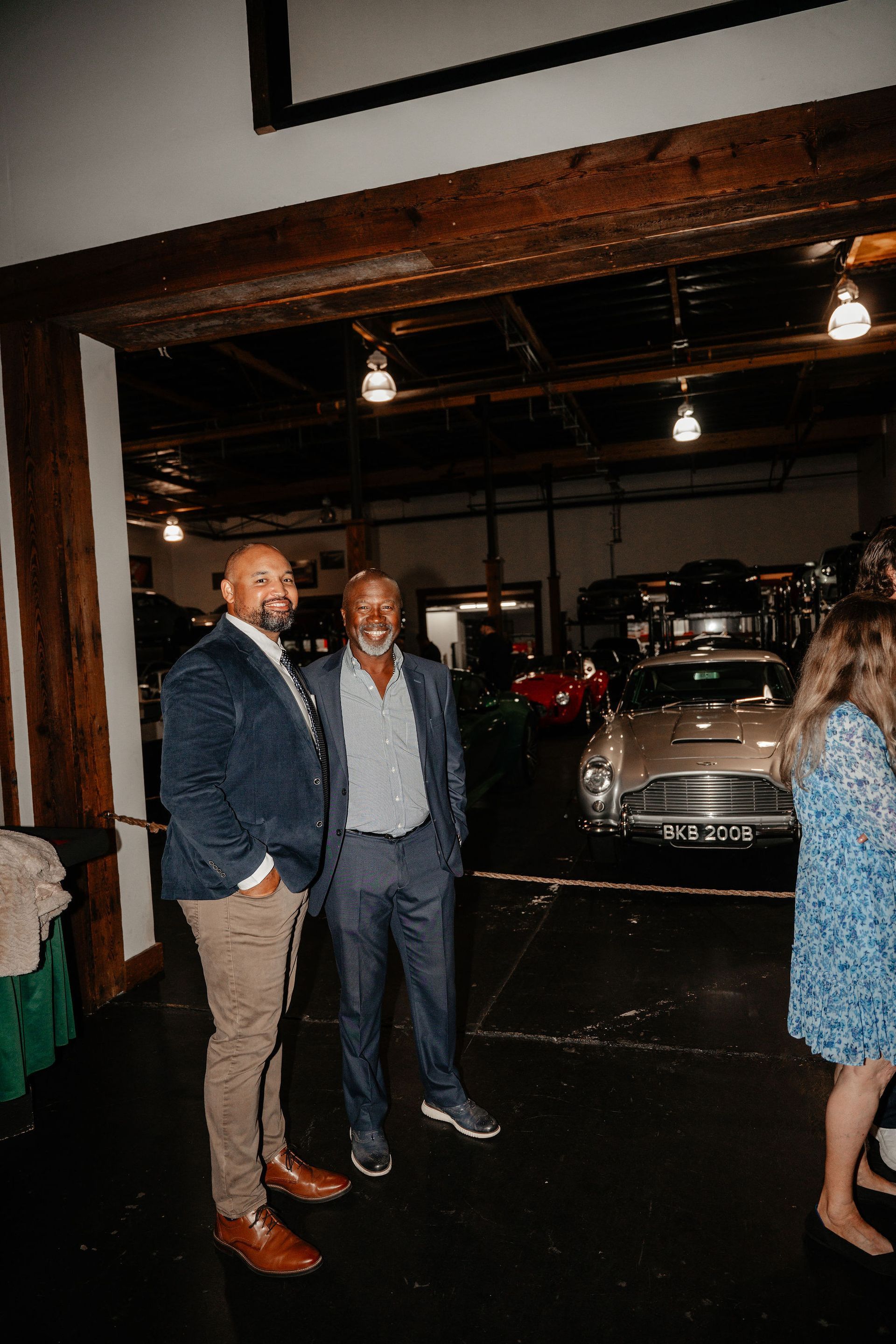 Two men pose in a dimly lit room with classic cars; one in a blue blazer and khakis, the other in a blue suit.