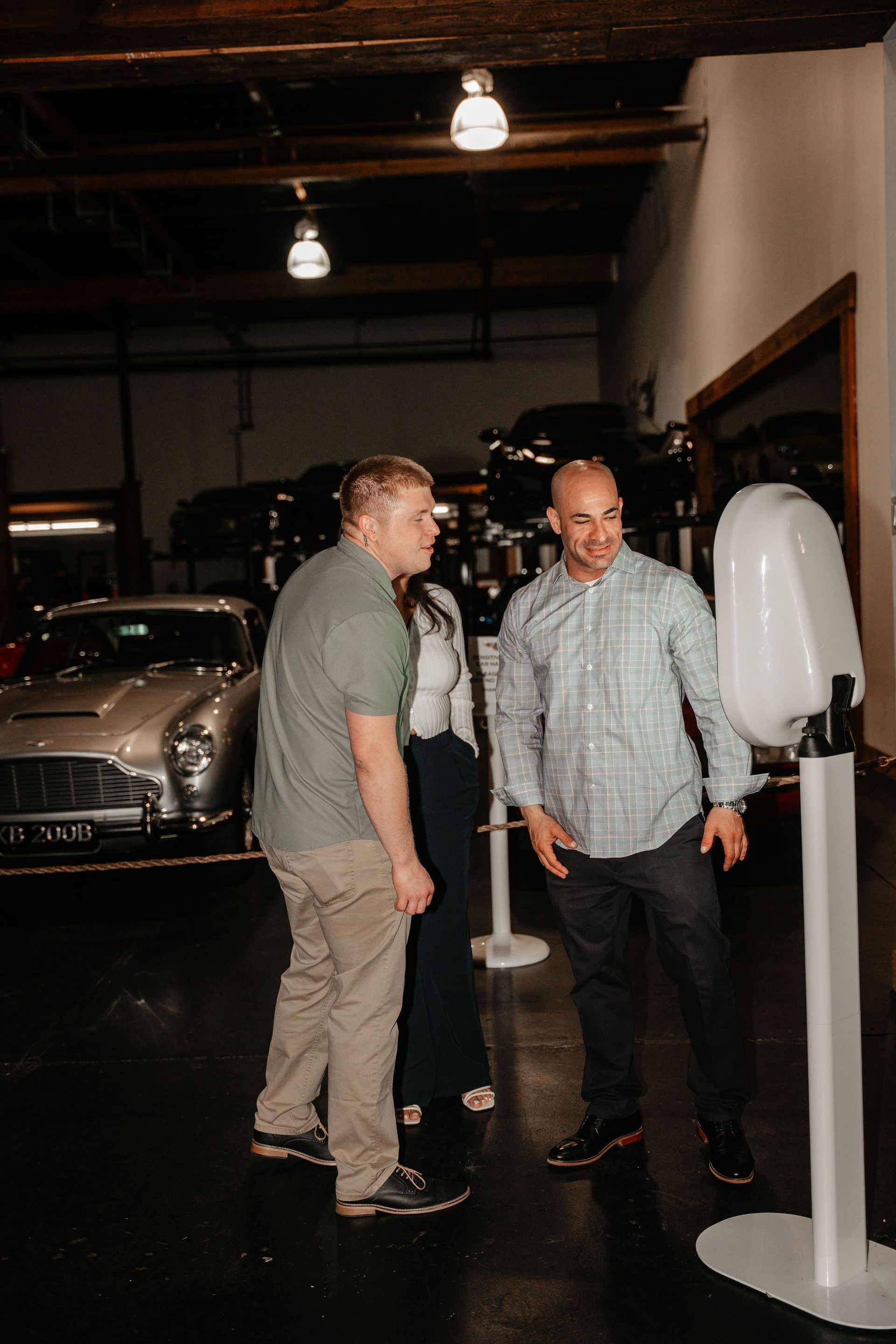 Two men and a woman by a photo booth in a garage with classic cars. The men are looking at the photo booth.