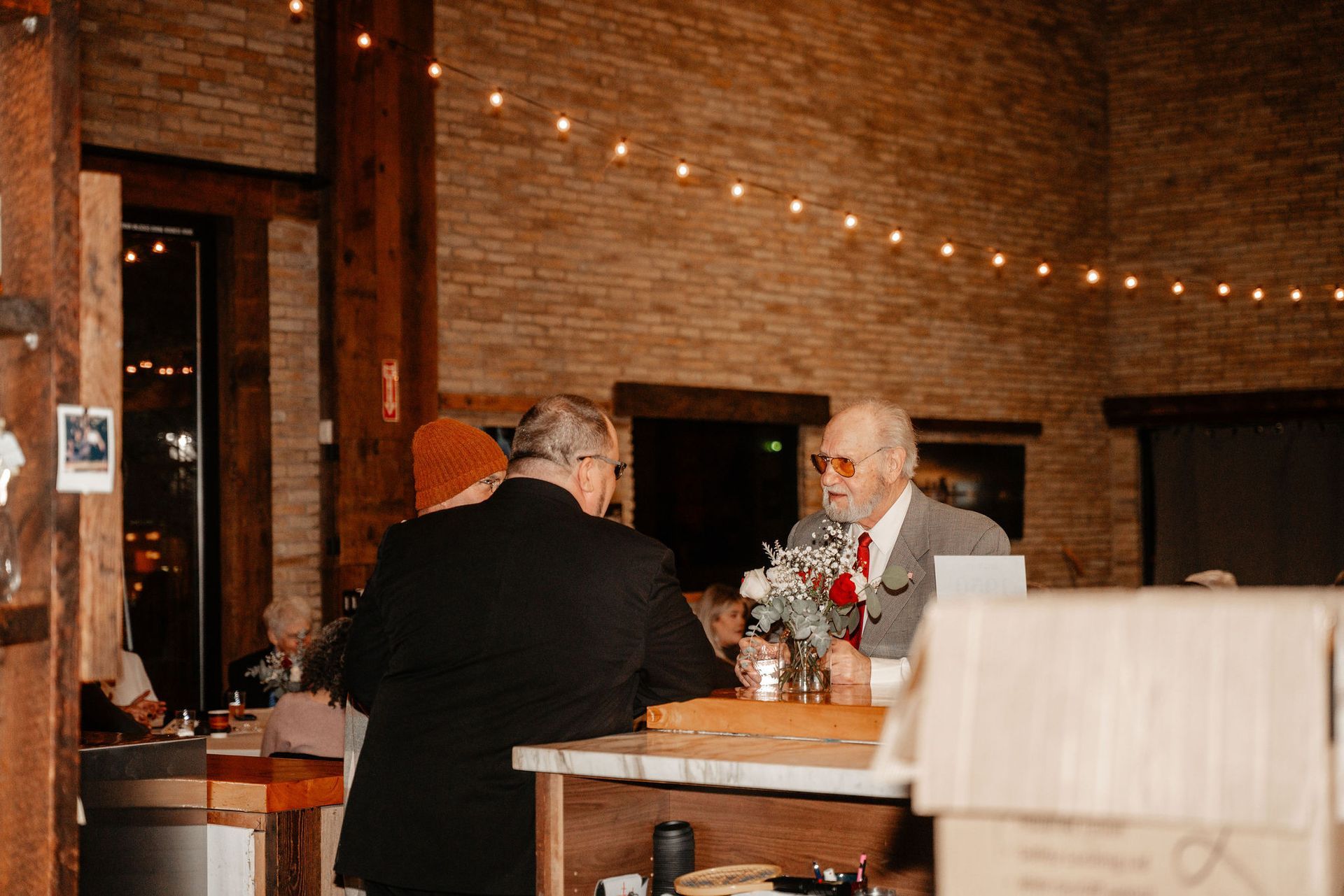 Two men in suits at a bar, talking, with a vase of flowers. Warm lighting in a brick building.