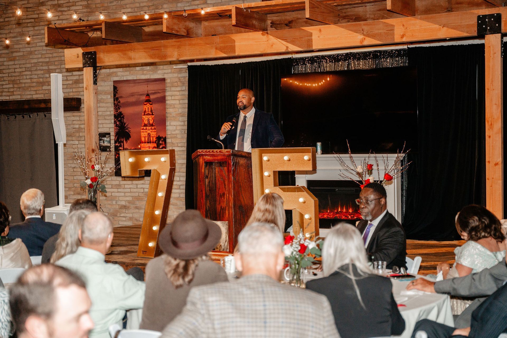 A man speaks at a podium at an event. Guests sit at tables with decorations, lit-up 