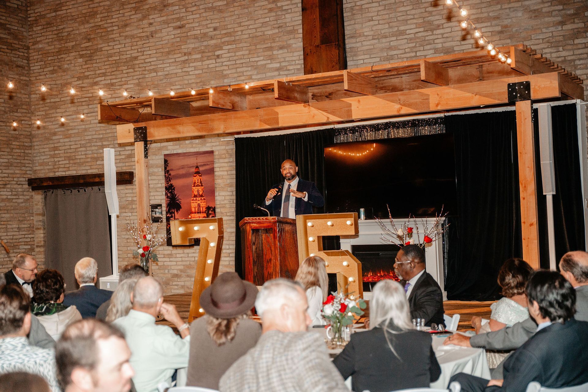 Man speaking at podium on stage, giving a presentation to seated audience. Stage has decorations.