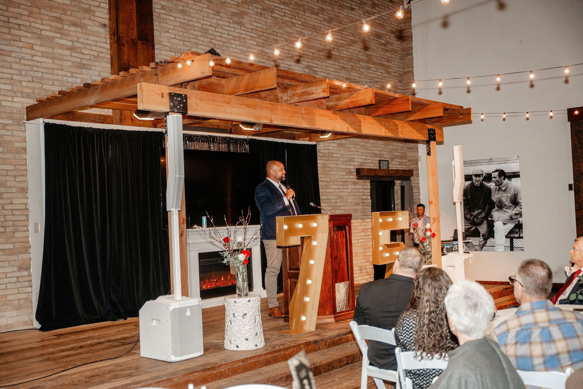 Man speaking at podium on stage with lit-up letters, under a wooden structure. Audience seated.