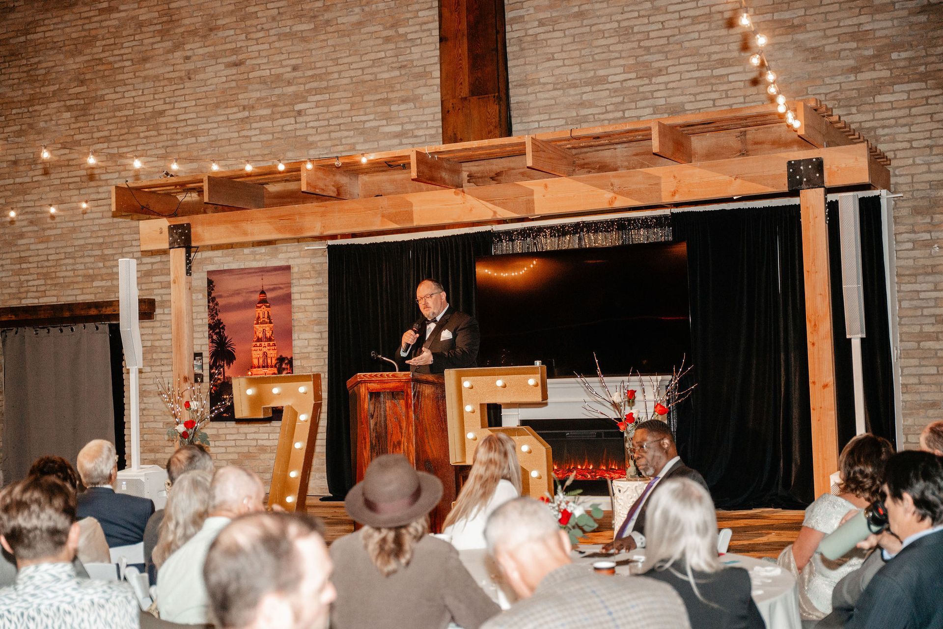 Man speaking at a podium on a stage with a wooden frame, and a crowd seated in an indoor setting.