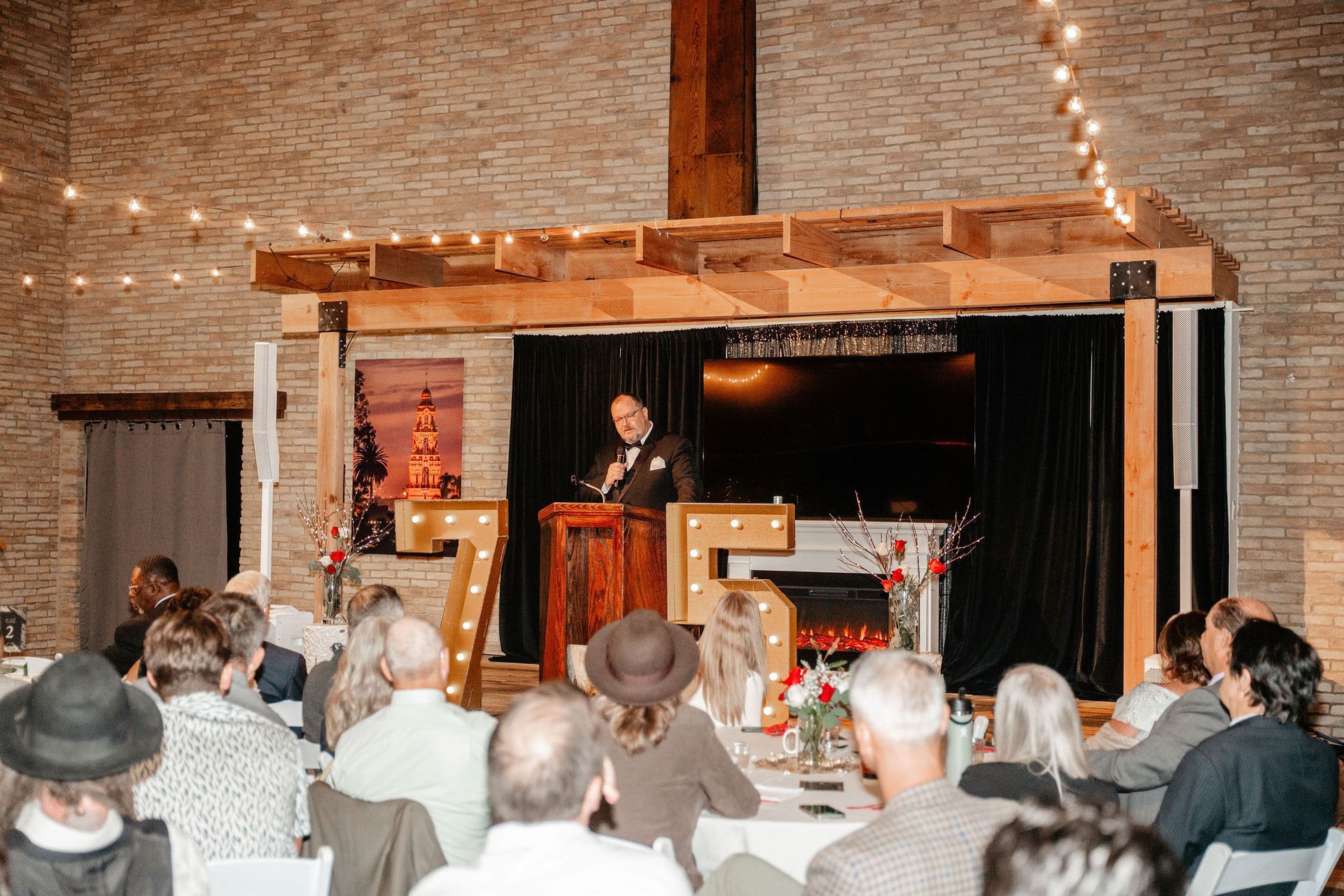 Man speaking at a podium in front of an audience. Backdrop with lit numbers, brick wall, and string lights.