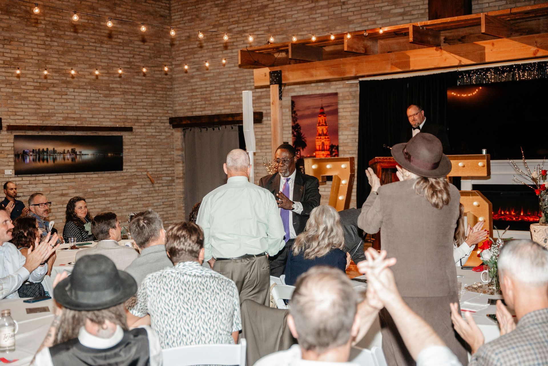 People clapping, celebrating at a gathering in a brick-walled room. A man speaks onstage.