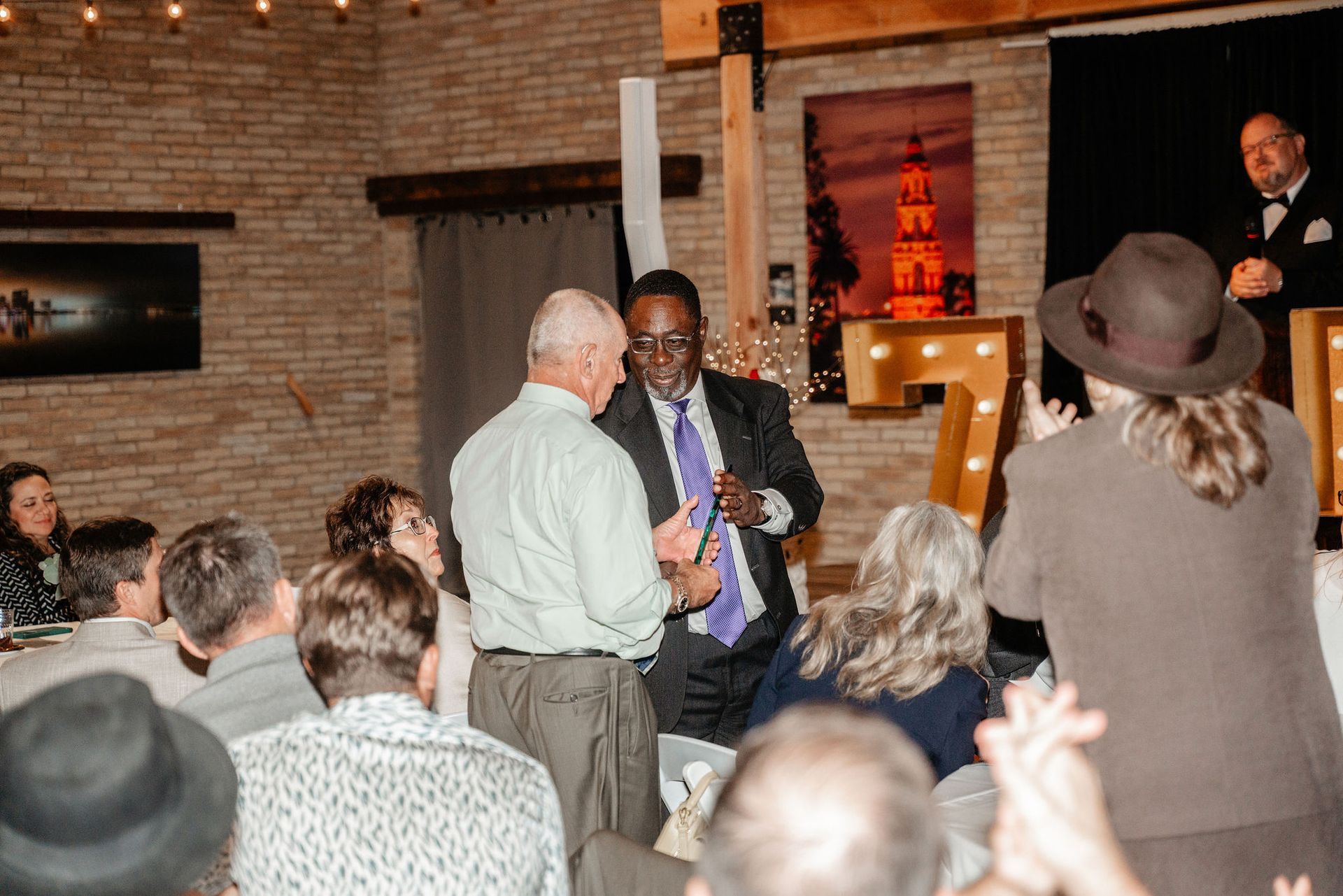 Man in suit receiving gift from man at event. Others applaud.  Brick wall and lit up letters in background.
