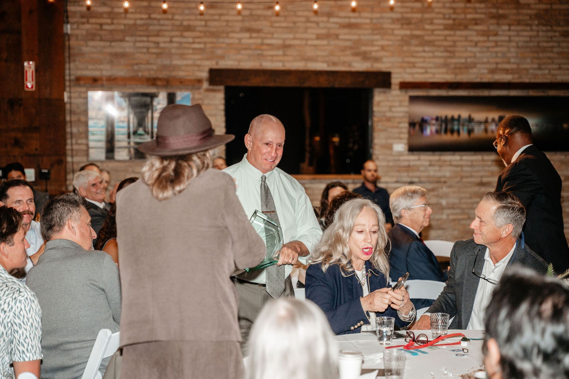 People at a gathering; woman in hat pulling a rope, man in light green shirt holding it, others seated at tables.