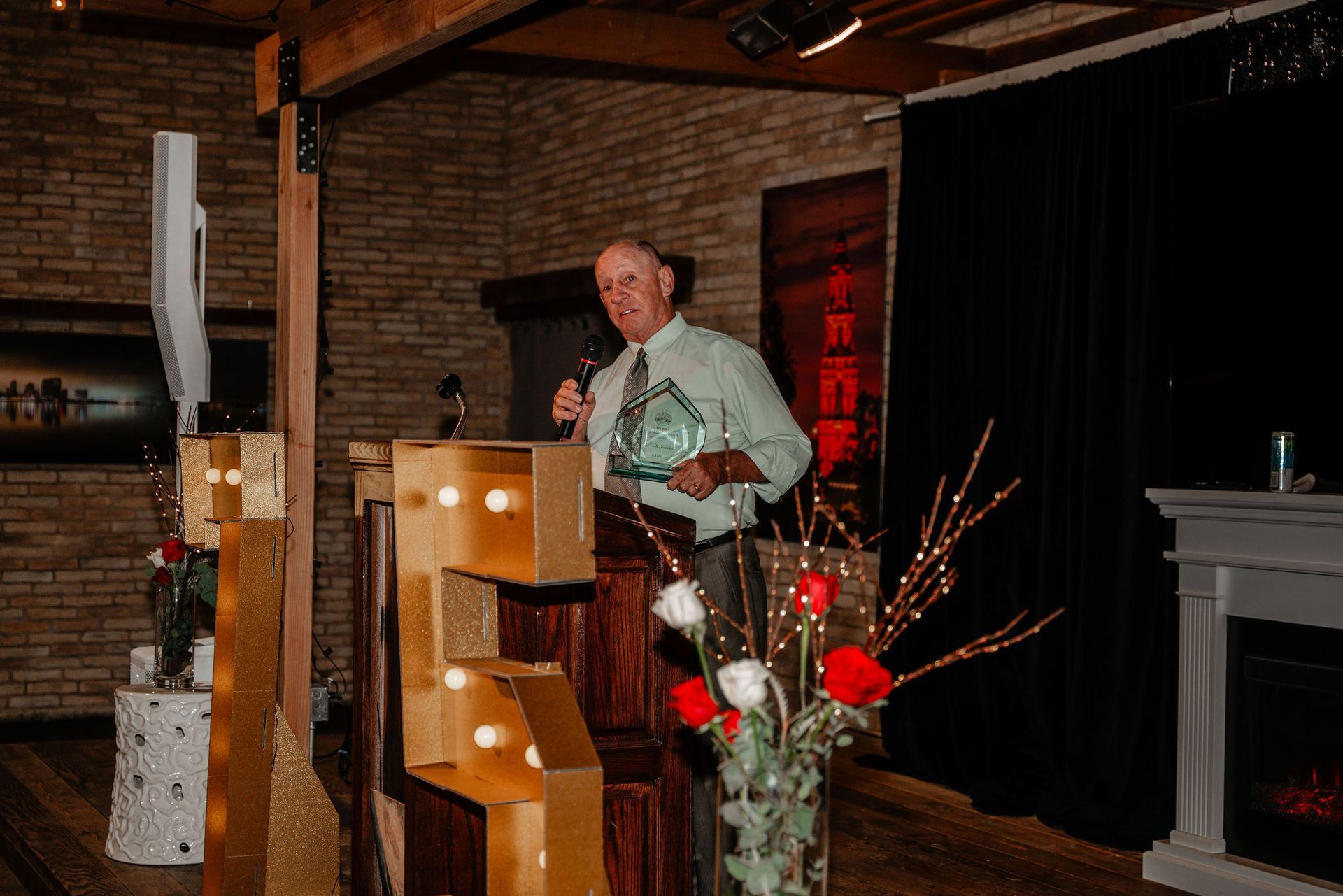 Man holding award at podium, speaking. Brick wall backdrop, decorated with lights and flowers.