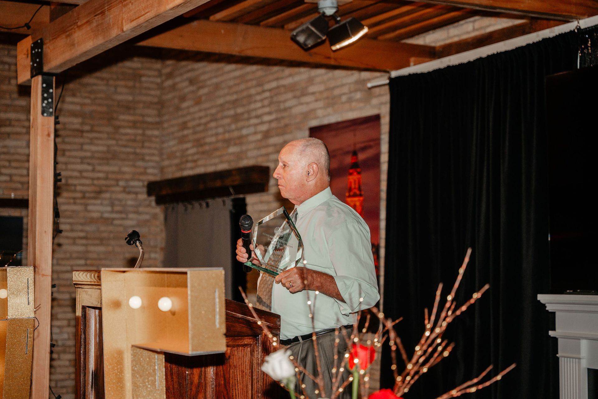 Man speaking at a podium in a brick-walled room, holding a microphone, with a dark backdrop.