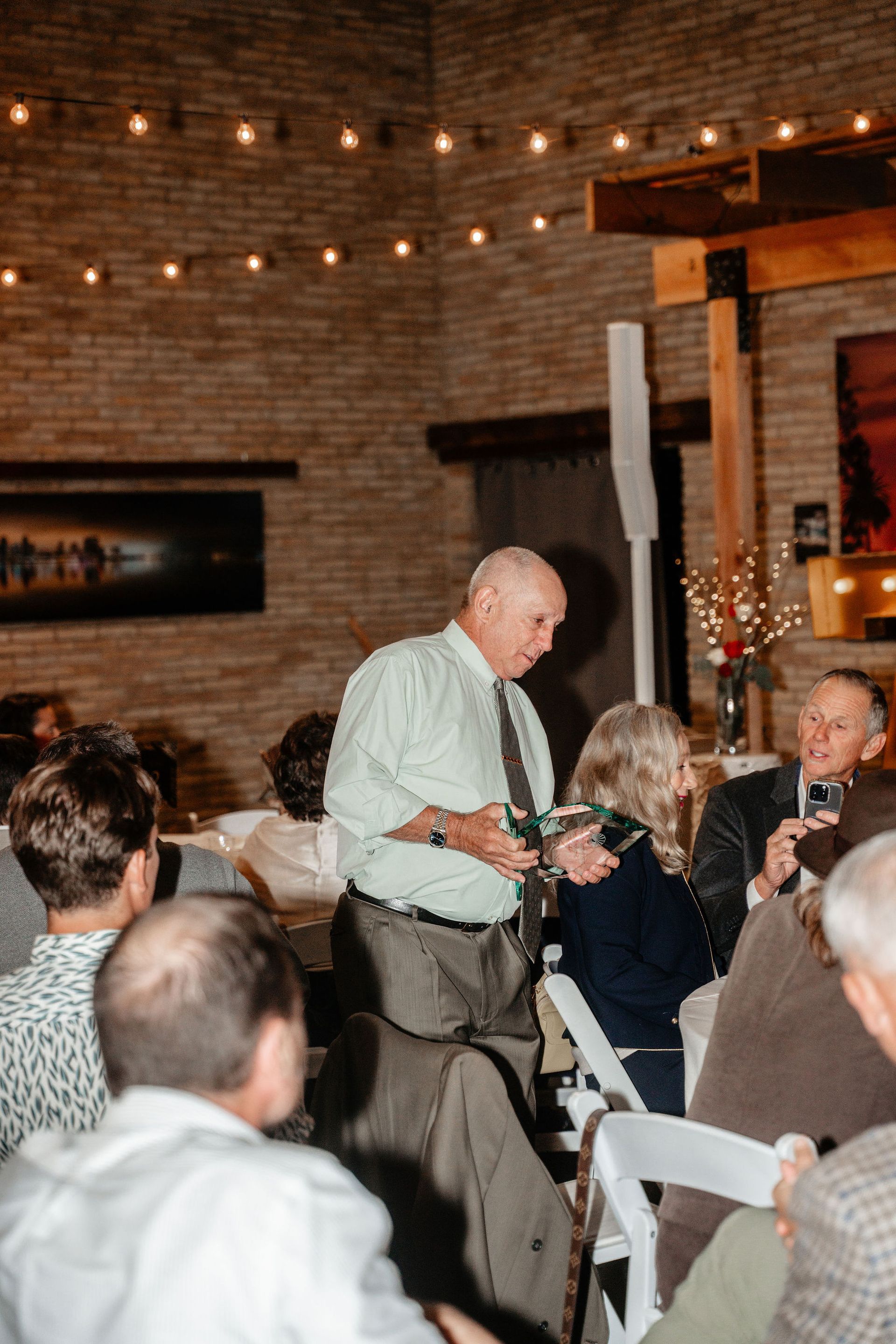 Man holding small object, speaking to seated guests at an event, brick wall background with lights.